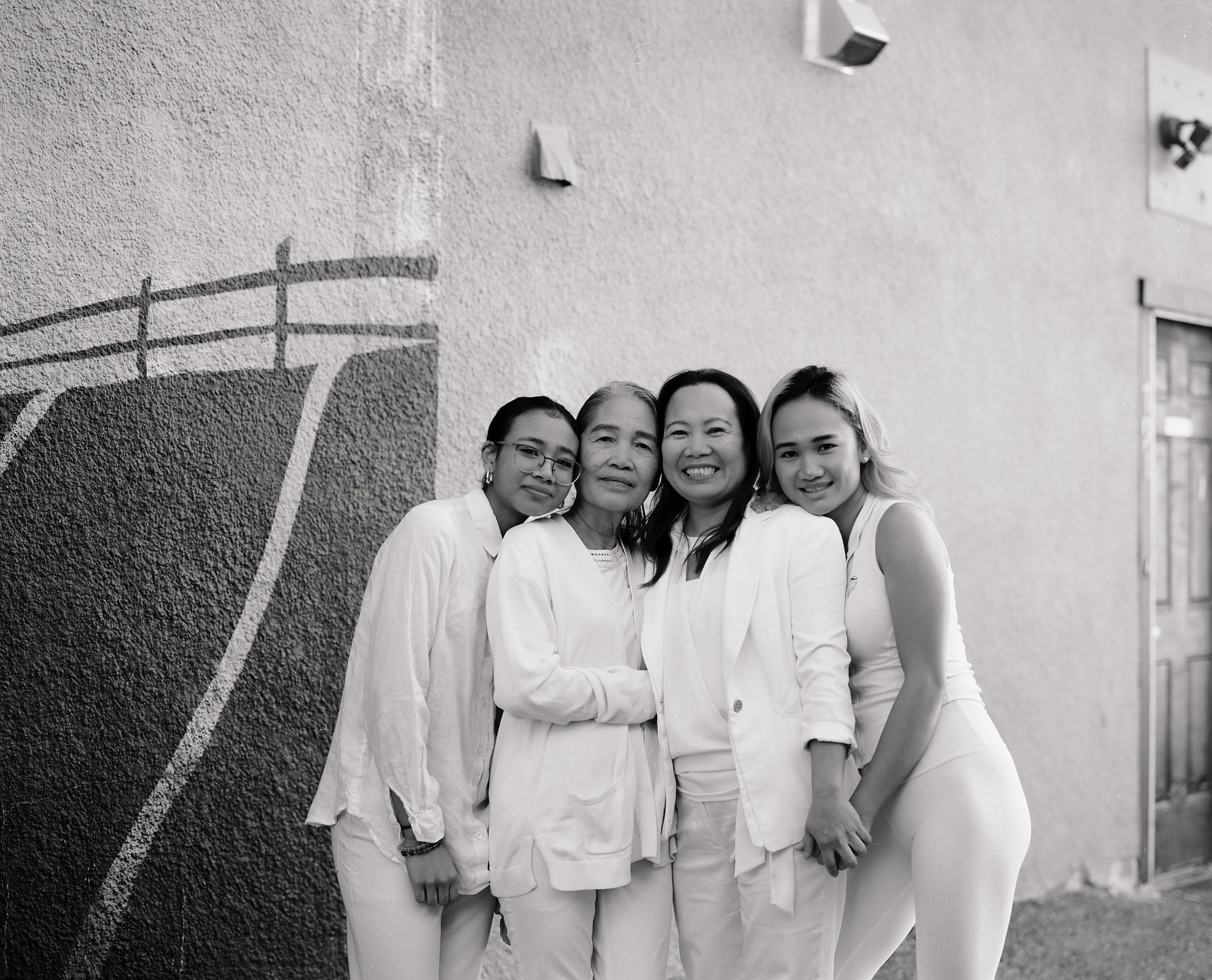 Four women standing together in front of a wall with a mural, smiling and embracing each other. The photo is in black and white.