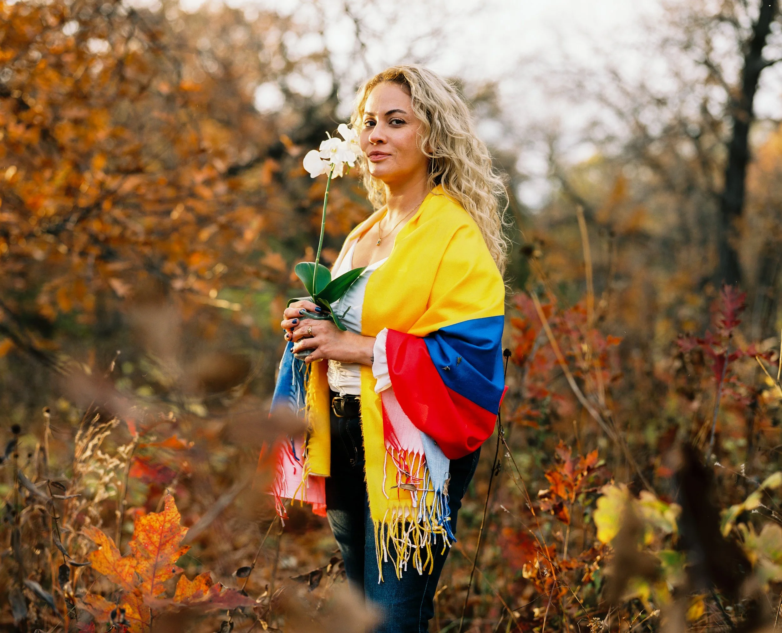 A woman with wavy blonde hair standing outdoors in an autumn forest, wrapped in a colorful flag with yellow, blue, and red sections, holding a white flower, and looking at the camera with a confident expression.