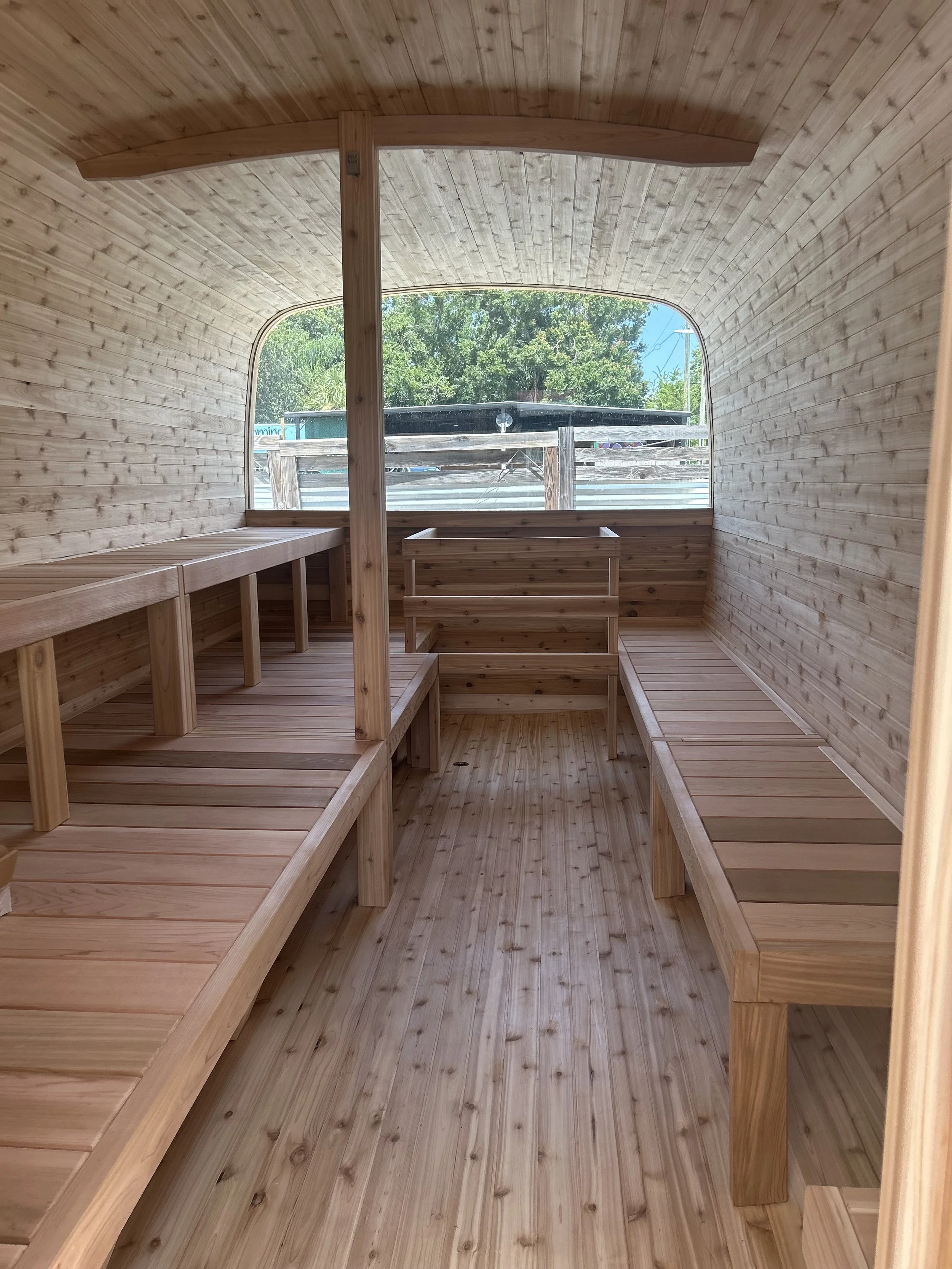The interior of a small wooden sauna with wooden benches on both sides and a large window showing outdoor trees and sky.