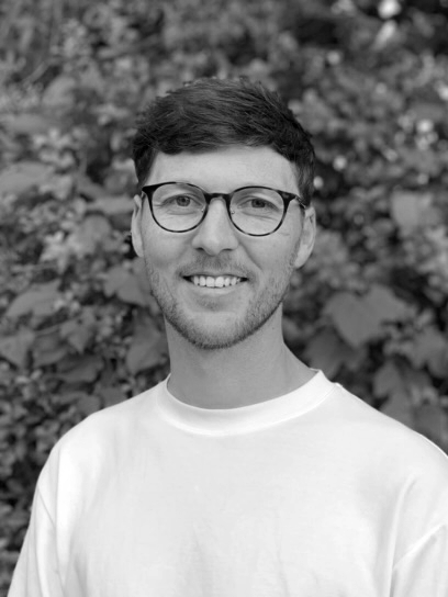 A black and white photo of a young man with glasses and a beard, smiling, standing outdoors in front of leafy background.