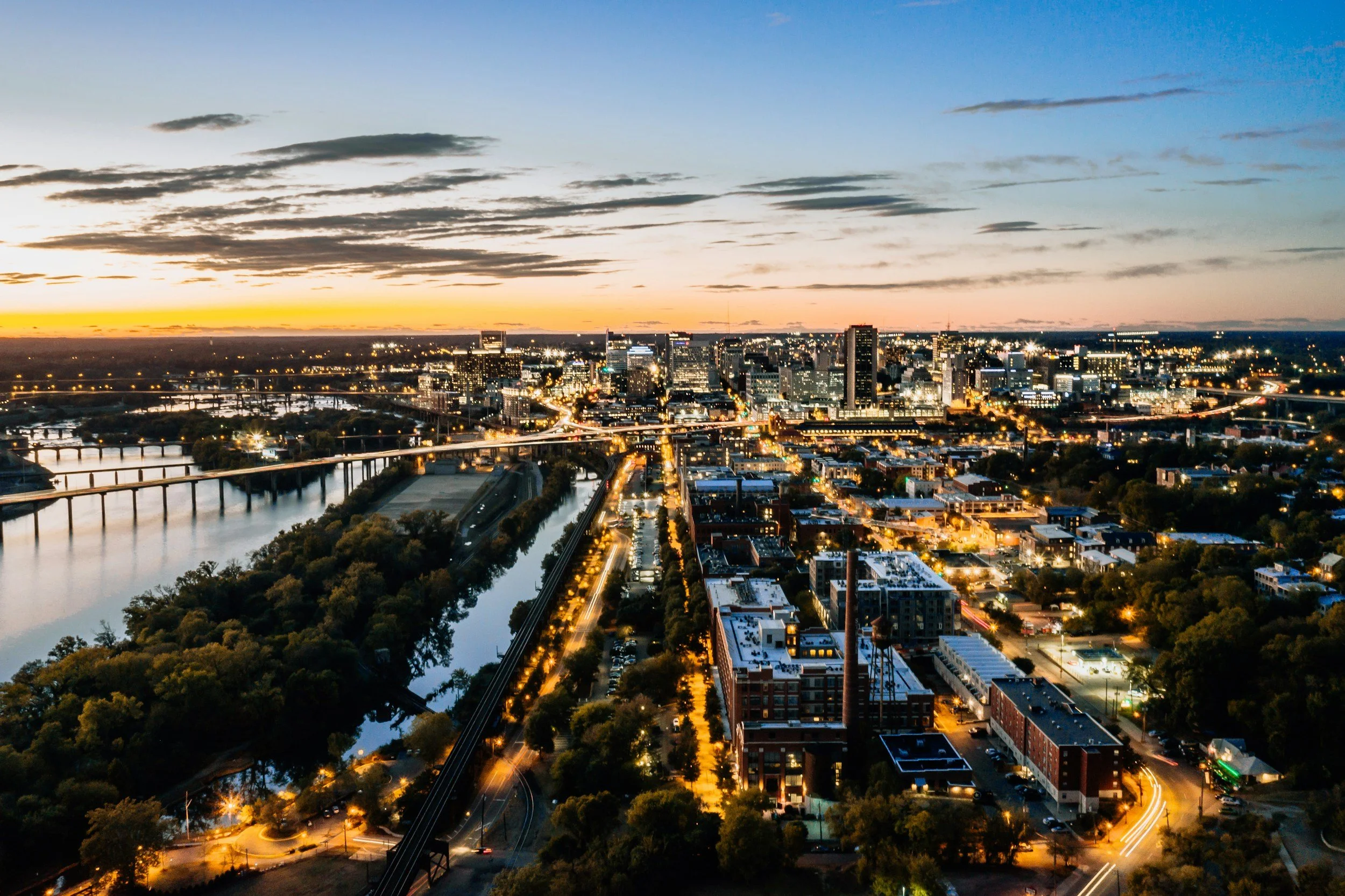 Aerial view of a city skyline at dusk with lit buildings, bridges over rivers, and streets illuminated by streetlights.