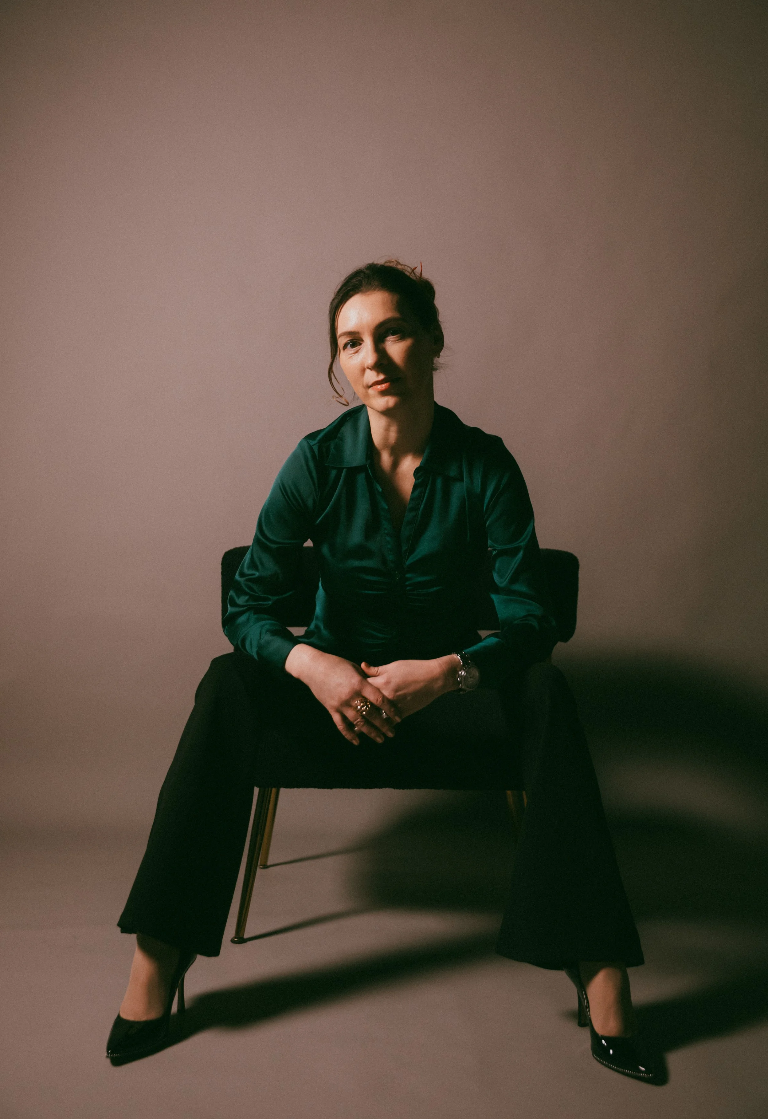 Dr. Kat sitting in a chair in a studio behind a warm brackground, wearing a teal blouse and black pants, leaning in her thighs and smiling at the camera