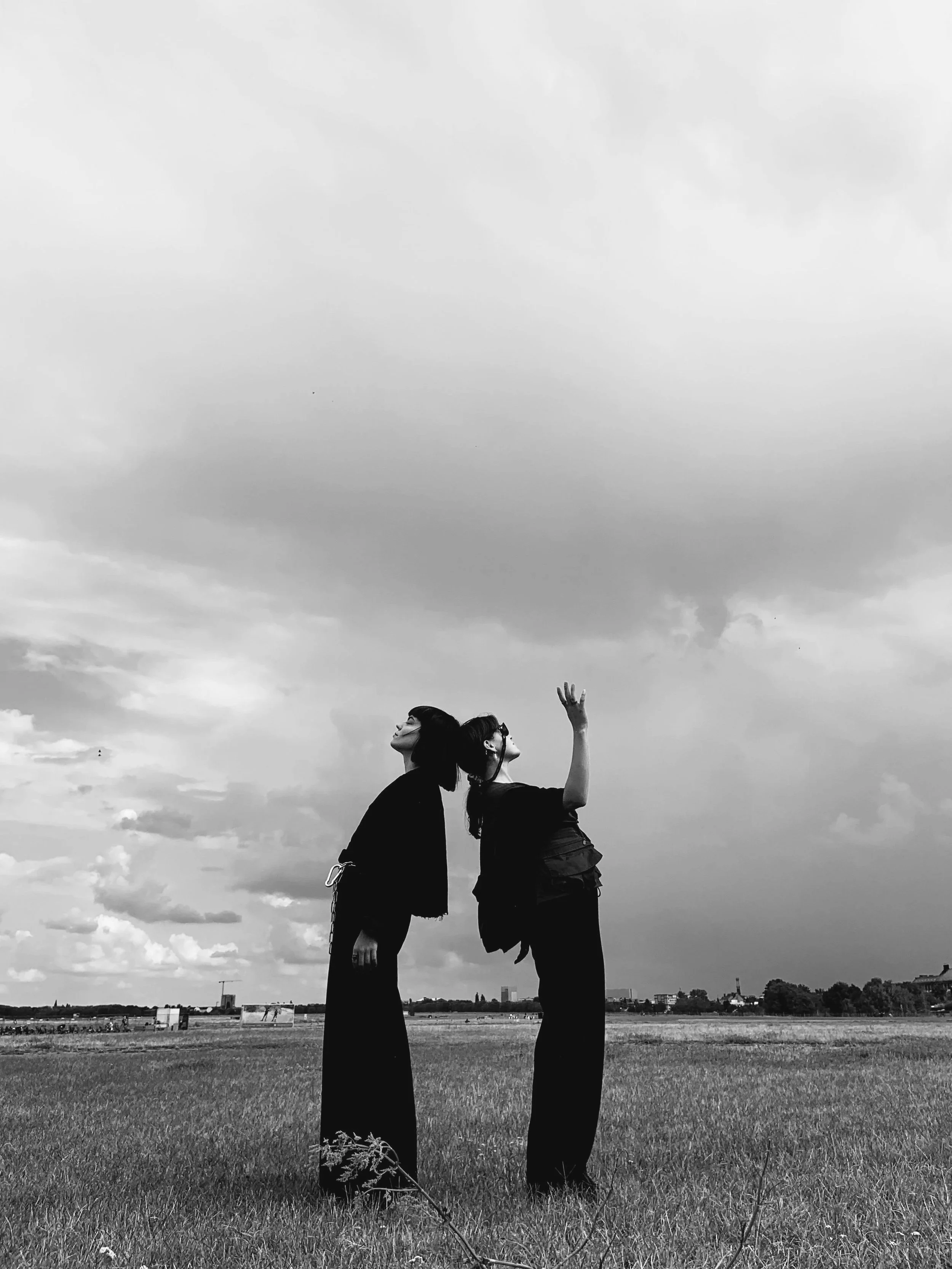 Two women in black outfits standing back-to-back in an open field under a dramatic sky, evoking strength, expansion, and transformation.