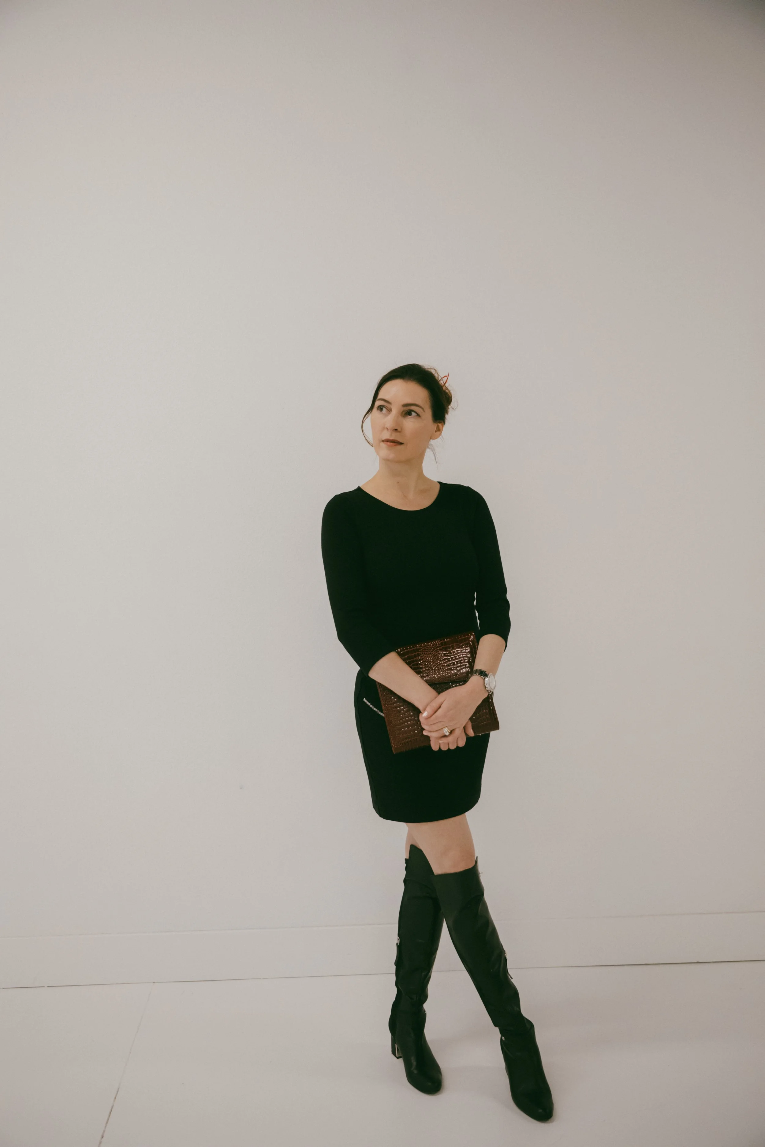 Dr. Kat in a studio with cream background, wearing a black dress and boots, holding a red leather planner in front of her, one legged crossed, and looking into the distant off camera