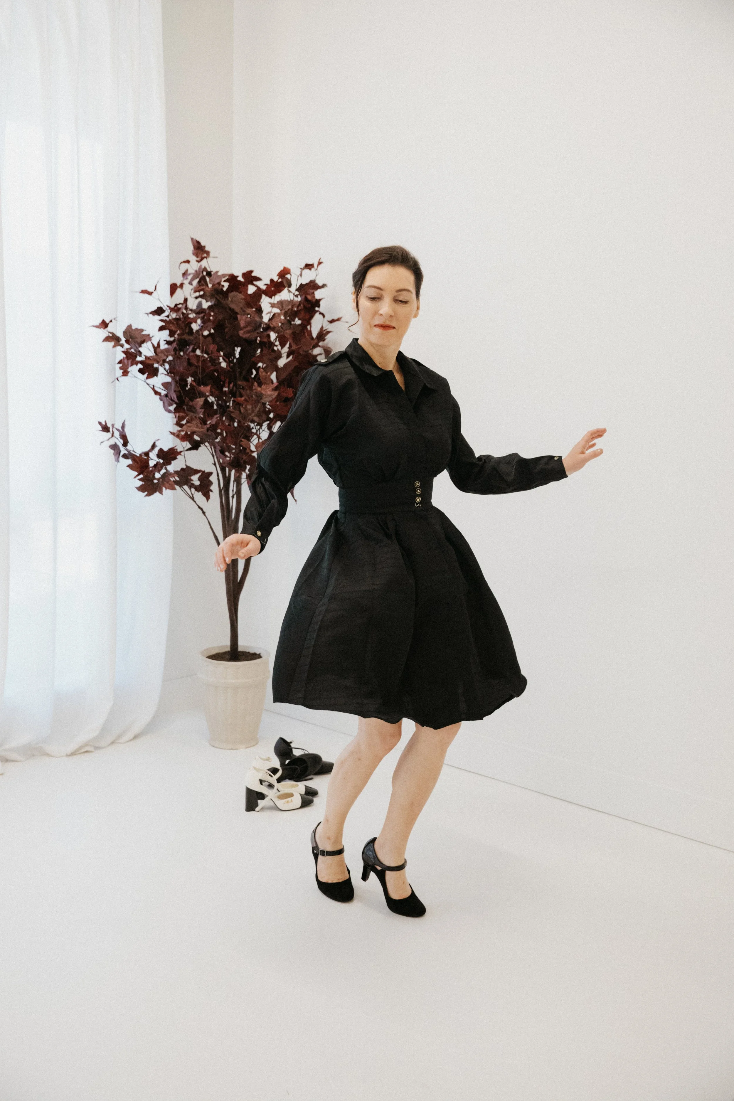 Dr. Kat wearing a black dress and black Mary Janes, in a dance pose, in a white studio, embodying movement and experimentation.