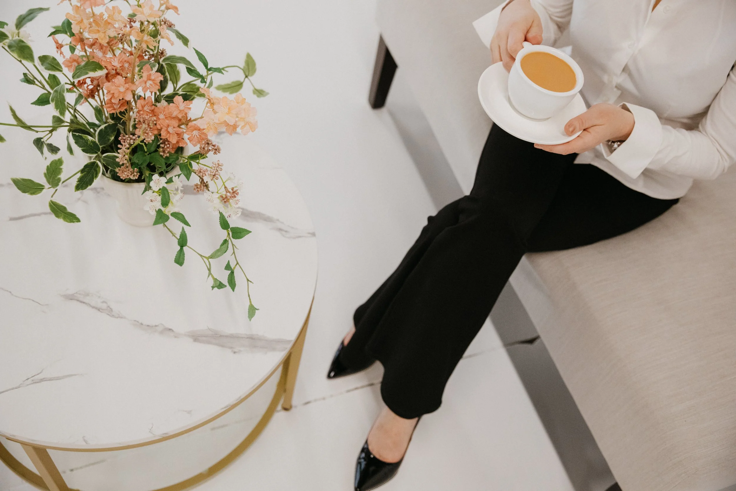 Overhead view of Dr. Kat seated with a cup of coffee beside a marble table with soft floral arrangement, reflecting a calm, refined, and thoughtful studio environment.