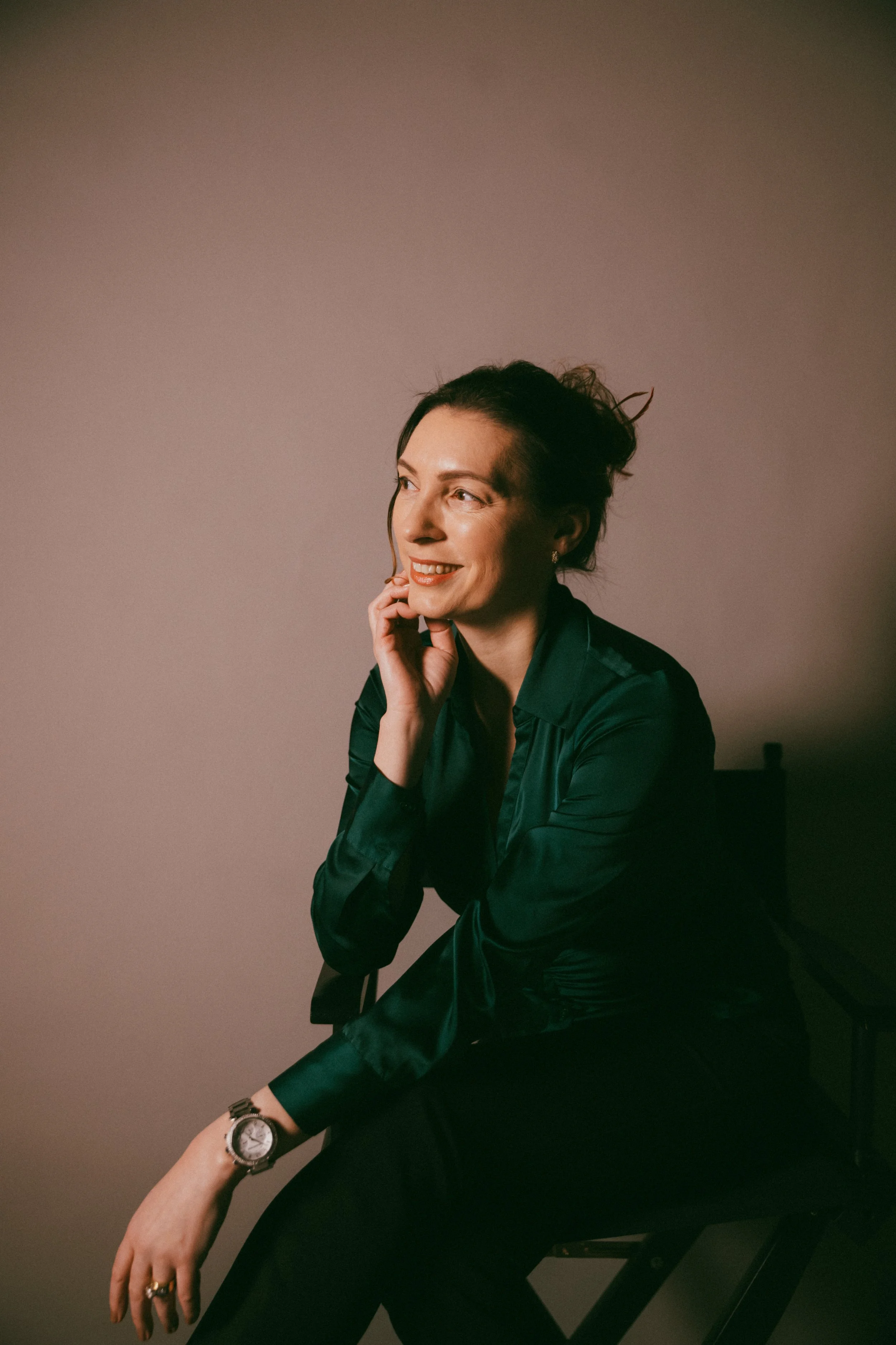 Dr. Kat in a study with warm grey background, sitting in a chair, hand on chin and smiling as in conversation, wearing a dark teal blouse and black trousers.