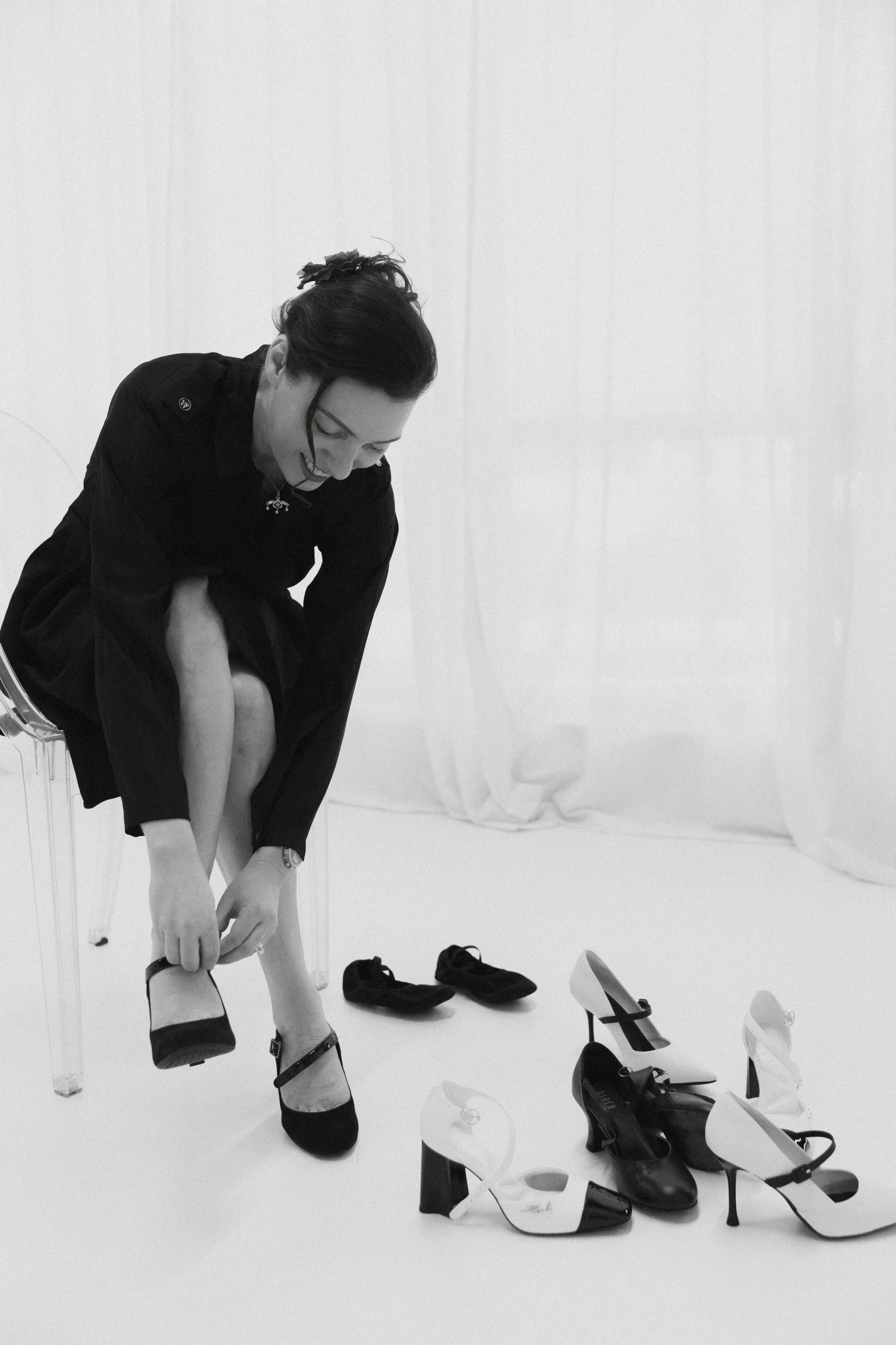 Dr. Kat seated in a softly lit studio adjusting her shoes beside several pairs of heels, reflecting personal transition, self-reinvention, and the process of finding stability inside growth and change.