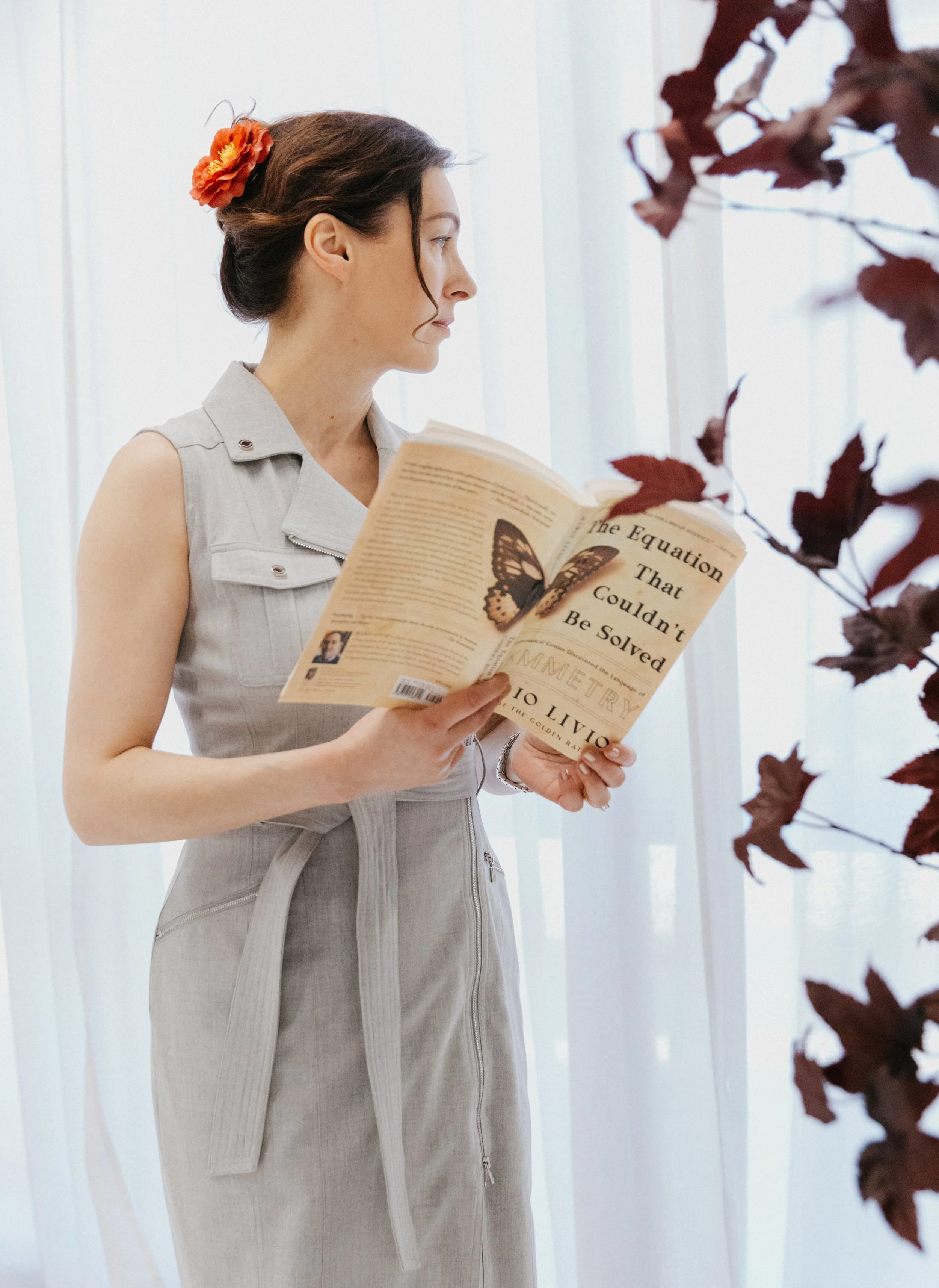 Dr. Kat standing thoughtfully with an open book in soft natural light, wearing a light gray dress beside deep red leaves in a calm studio setting that reflects curiosity, growth, and thoughtful self-development.