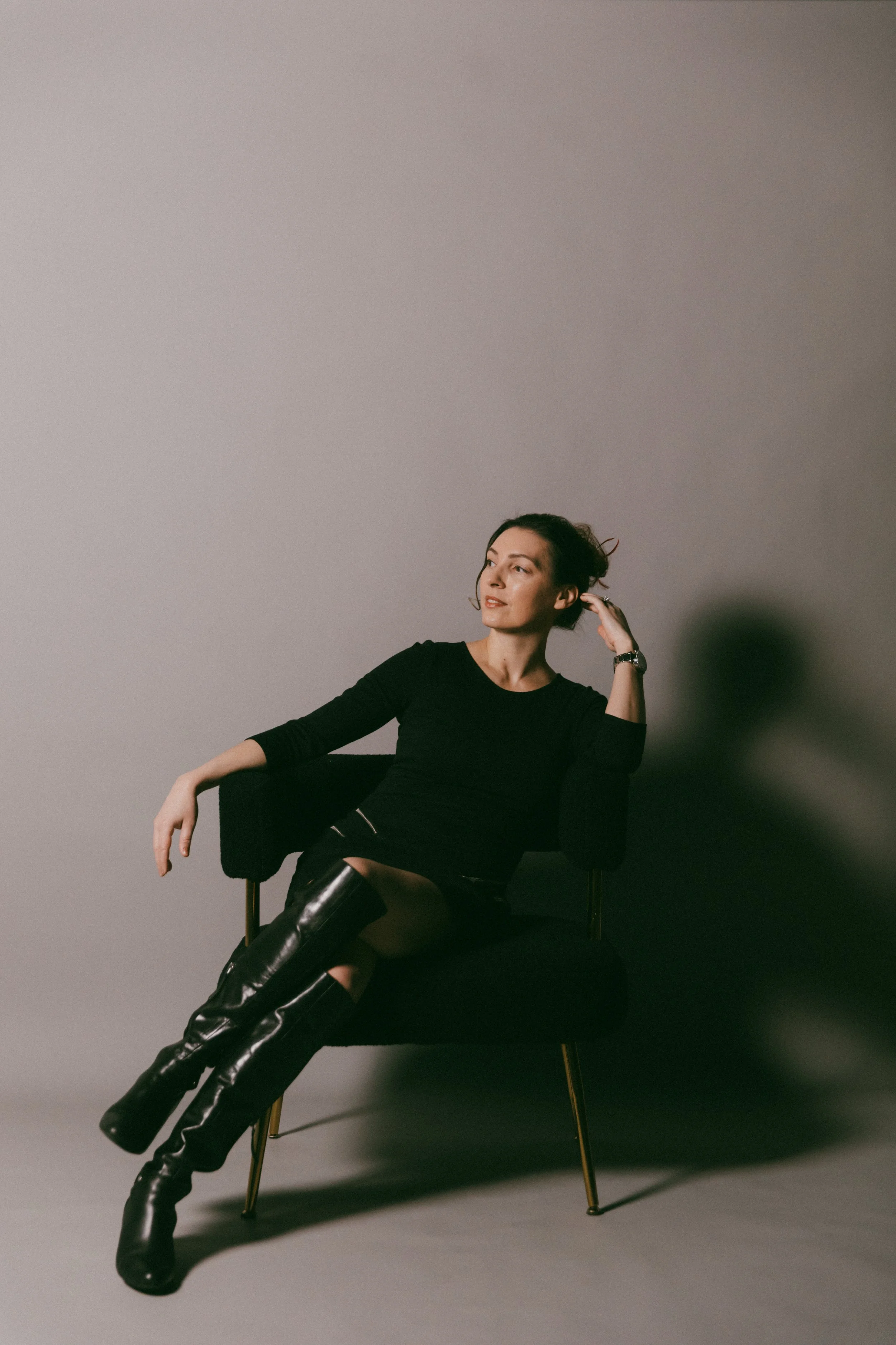 Dr. Kat sitting in a studio chair behind neutral background, wearing a black dress and boots, leaning thoughtfully in her hand and looking off to the side, smiling lightly