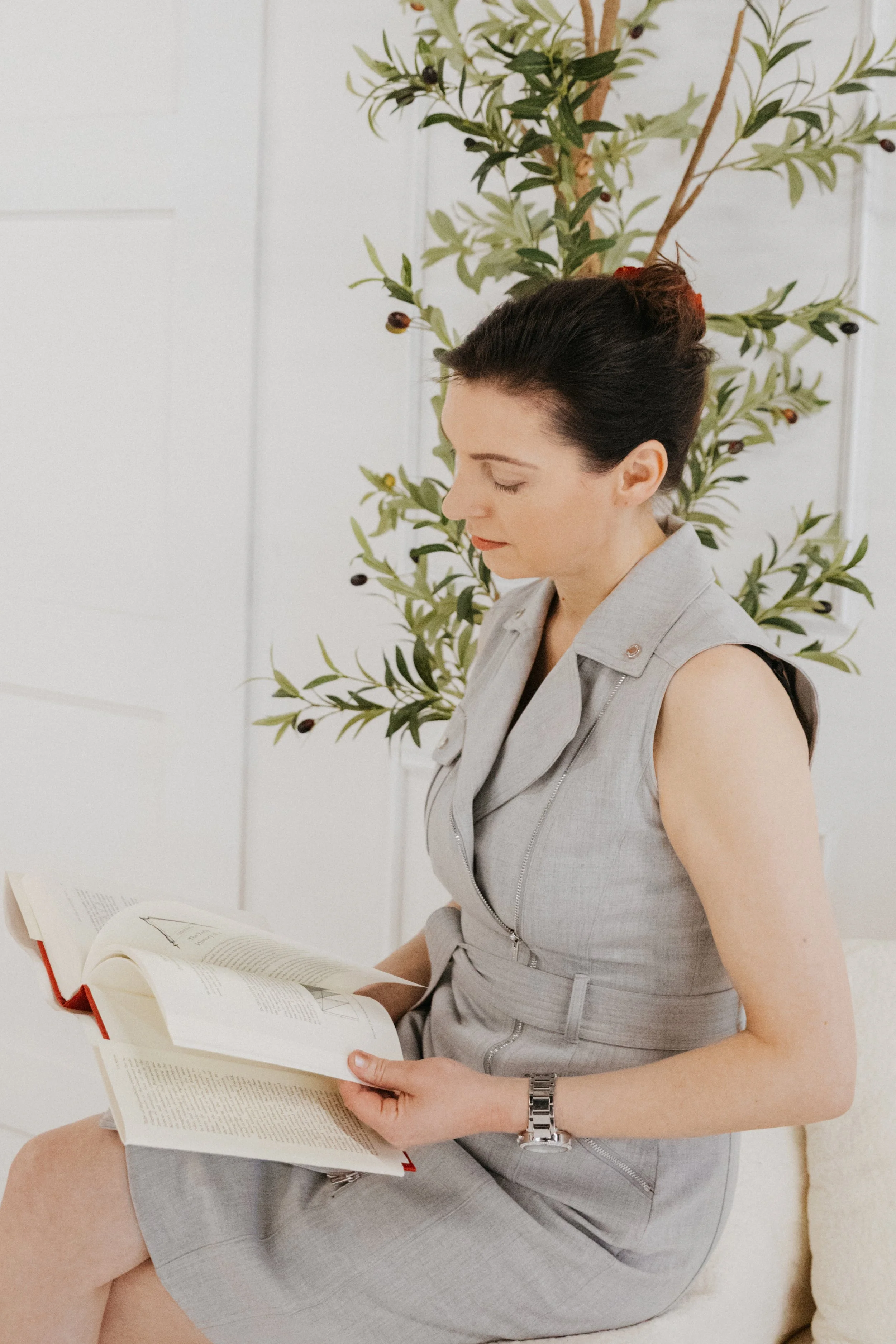 Dr. Kat seated with an open book in a soft, minimalist studio setting, wearing a light gray dress and reading thoughtfully beside a green olive tree in natural light.