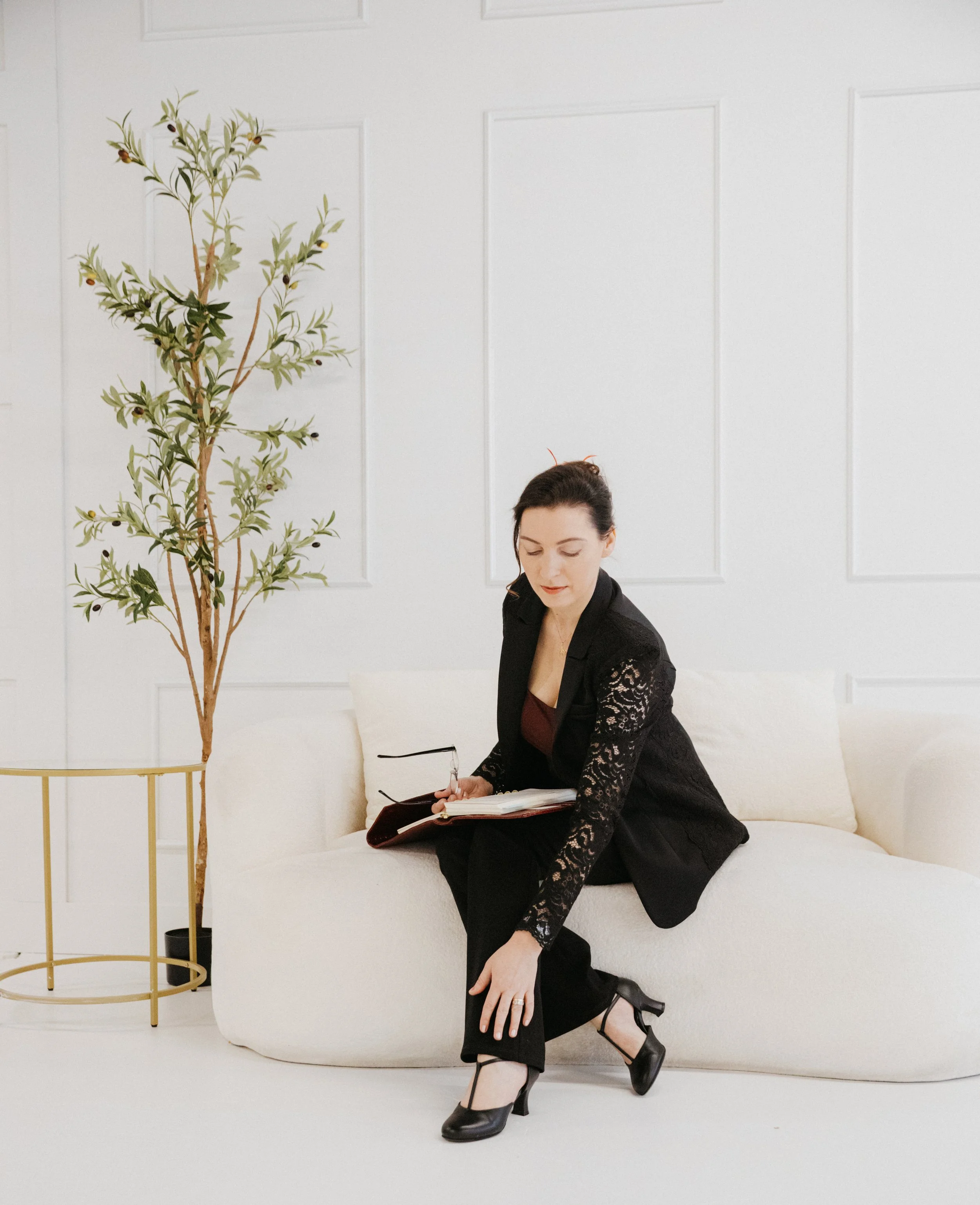Dr. Kat seated thoughtfully on a white sofa in a calm studio setting with a notebook in hand, reflecting Biological Leadership™, nervous system awareness, emotional presence