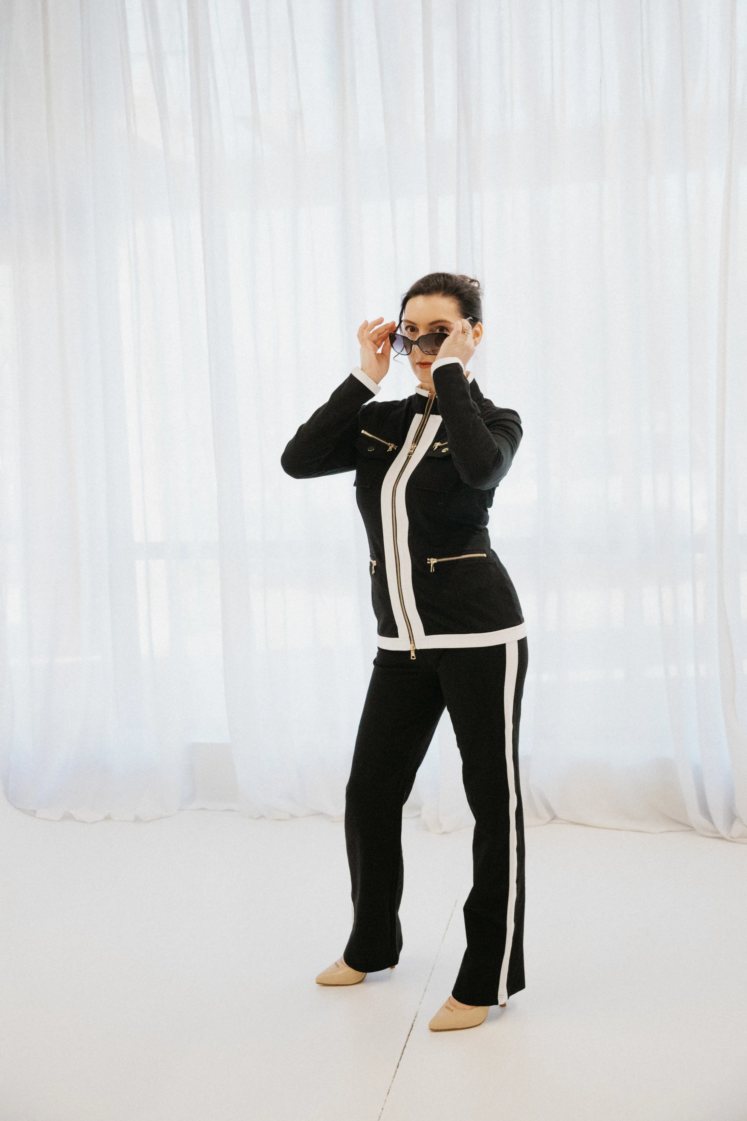 Dr. Kat wearing a black and white pant suit outfit, standing in front of a large window covered by white flowy curtains, holding the sides of her sunglasses like she is putting them on and peering over the glasses rims