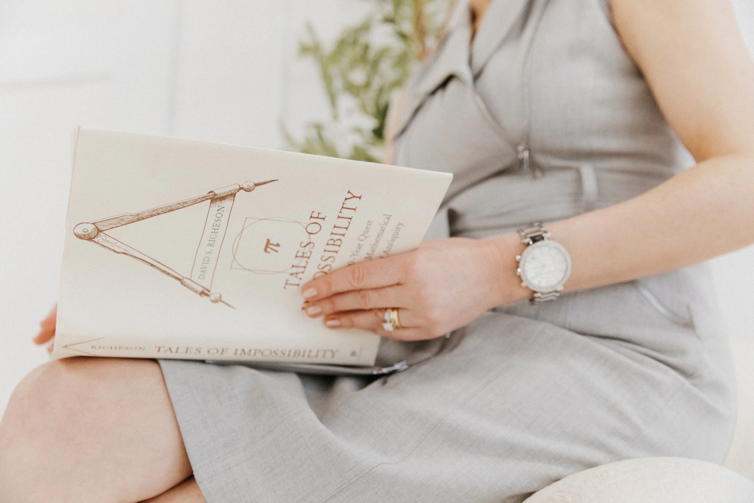 Close-up of Dr. Kat seated with a book in hand, wearing a light gray dress in a soft, minimalist studio setting that reflects thoughtful presence, learning, and quiet self-reflection.