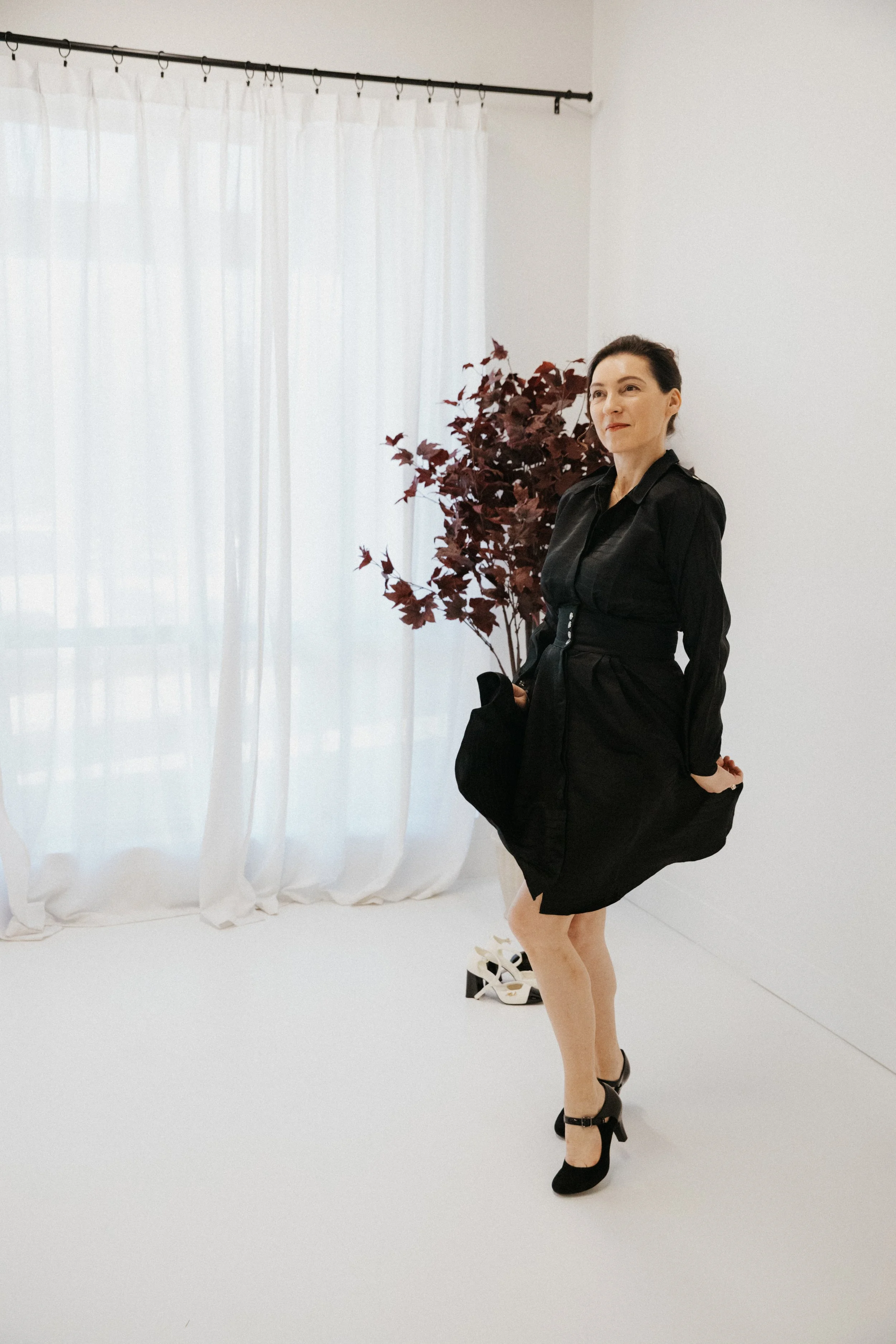 Dr. Kat standing calmly in a bright minimalist studio wearing a flowing black dress, reflecting ease, emotional safety, grounded confidence, and the feeling of becoming more fully yourself again.