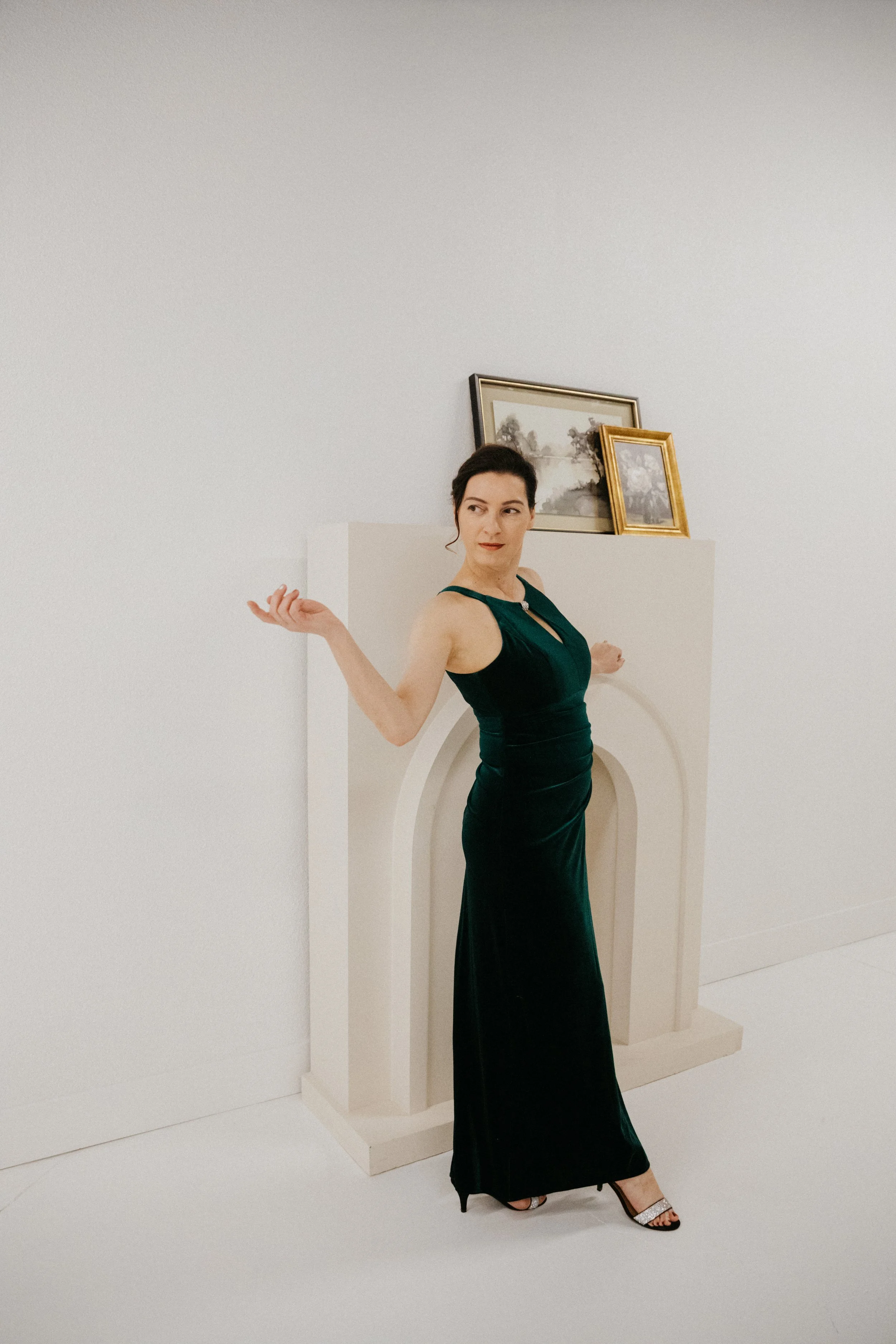 Dr. Kat standing in front of a cream fireplace with 2 paintings on the mantel, she wears a green velvet evening dress and black sandals, looking over her shoulder and her right arm extended