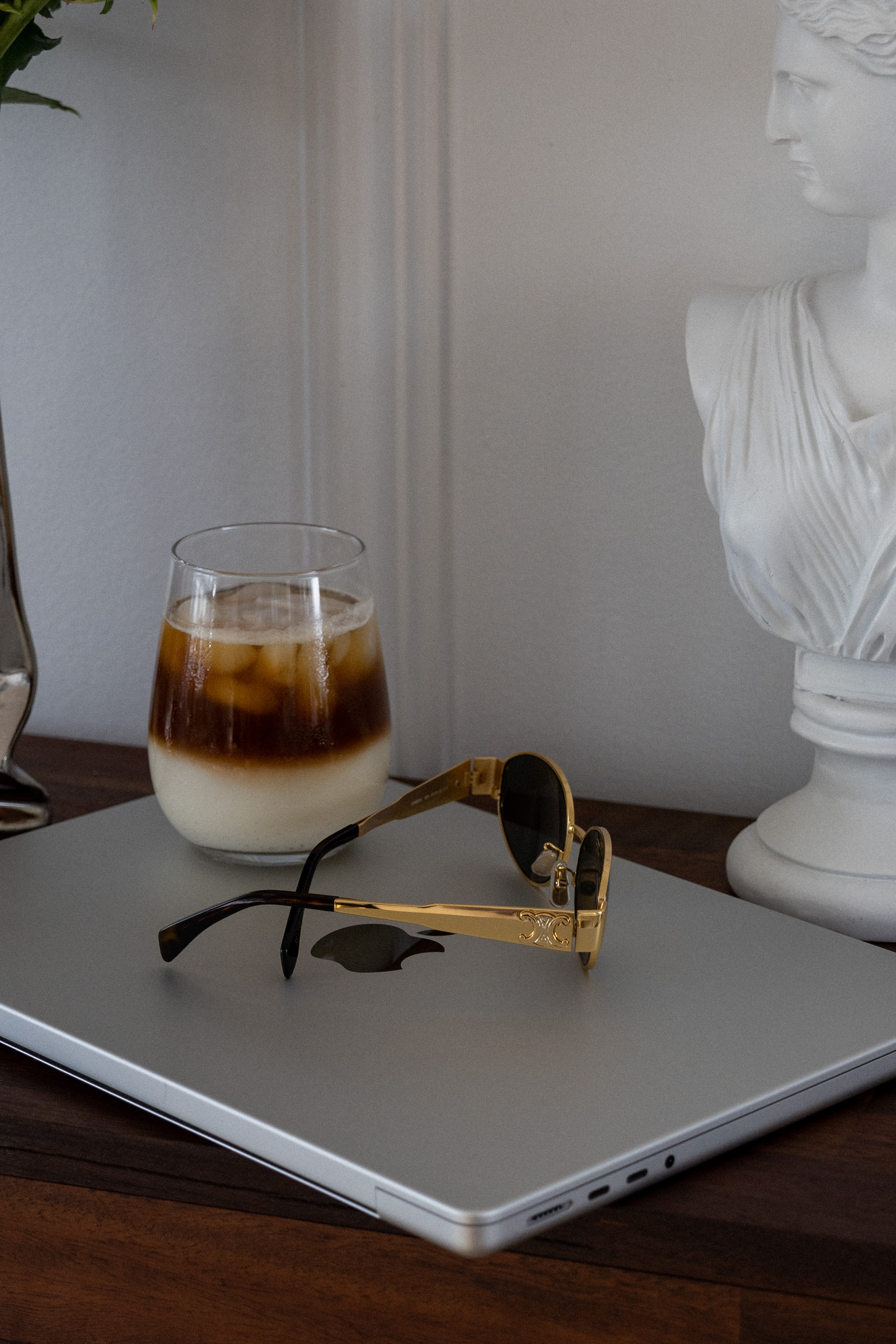 Minimalist workspace with iced coffee on a laptop, sunglasses, and classical sculpture—representing focused work, calm energy, and intentional capacity for high-performing professionals.