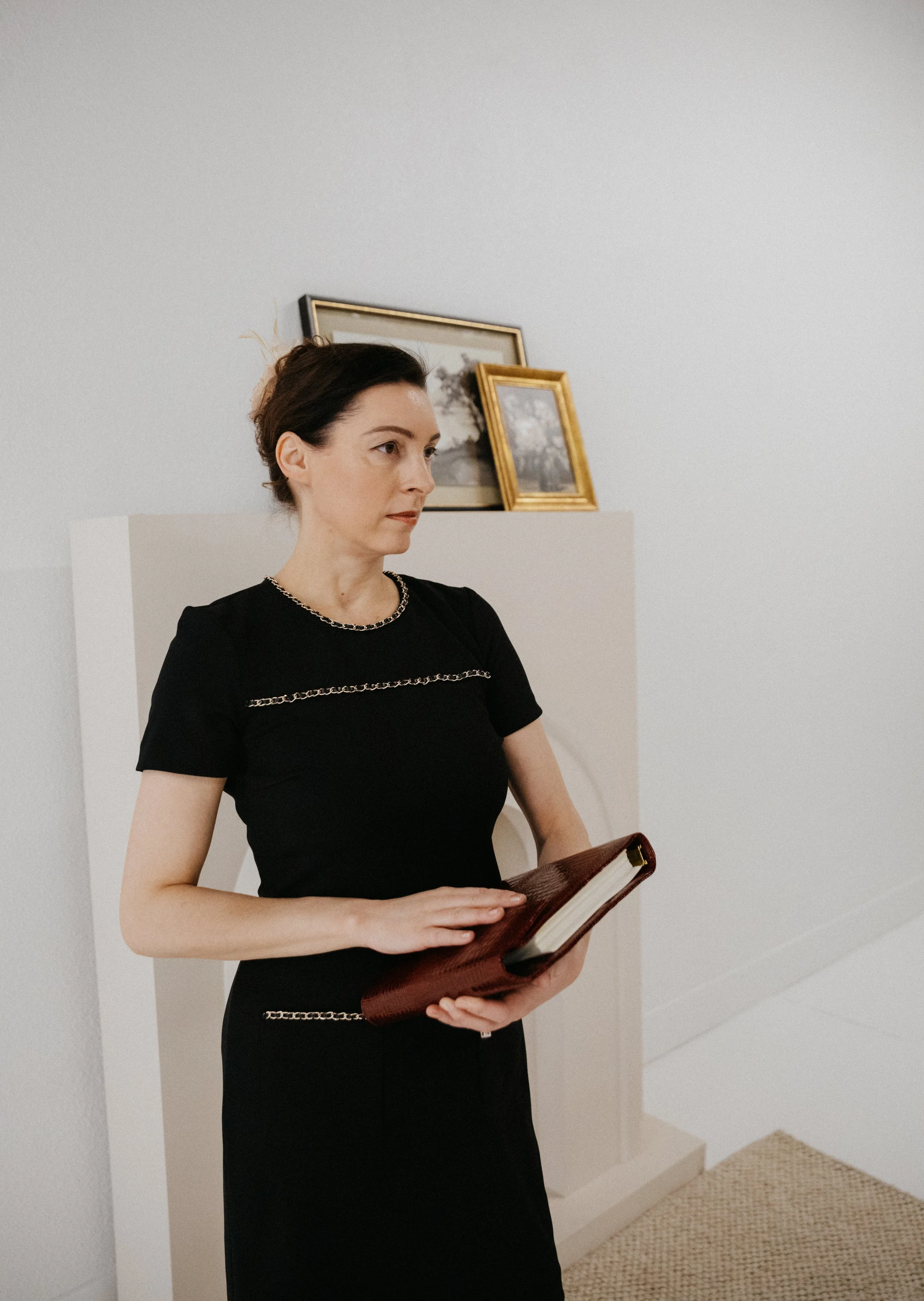 Dr. Kat standing in a minimalist interior holding a leather notebook, wearing a black dress and reflecting a calm, thoughtful, and grounded presence.