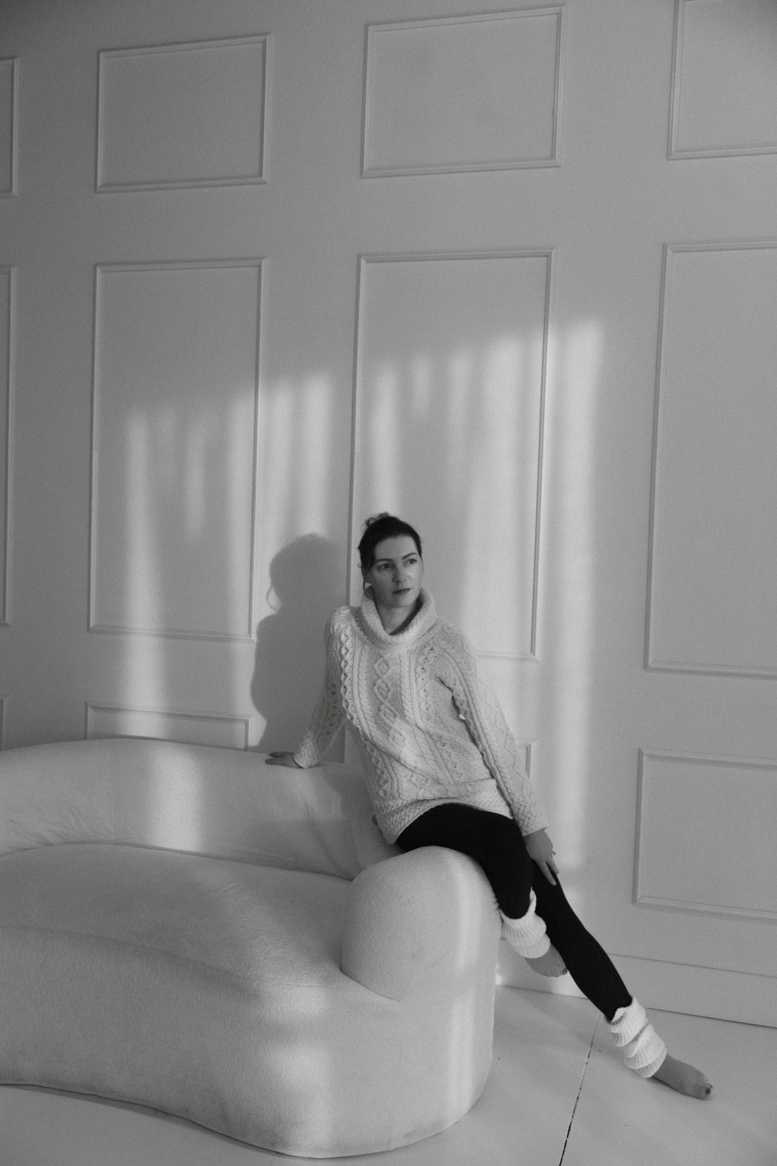 A black and white image of Dr. Kat in a studio, sitting on the edge of a sofa and looking thoughtfully into the distance