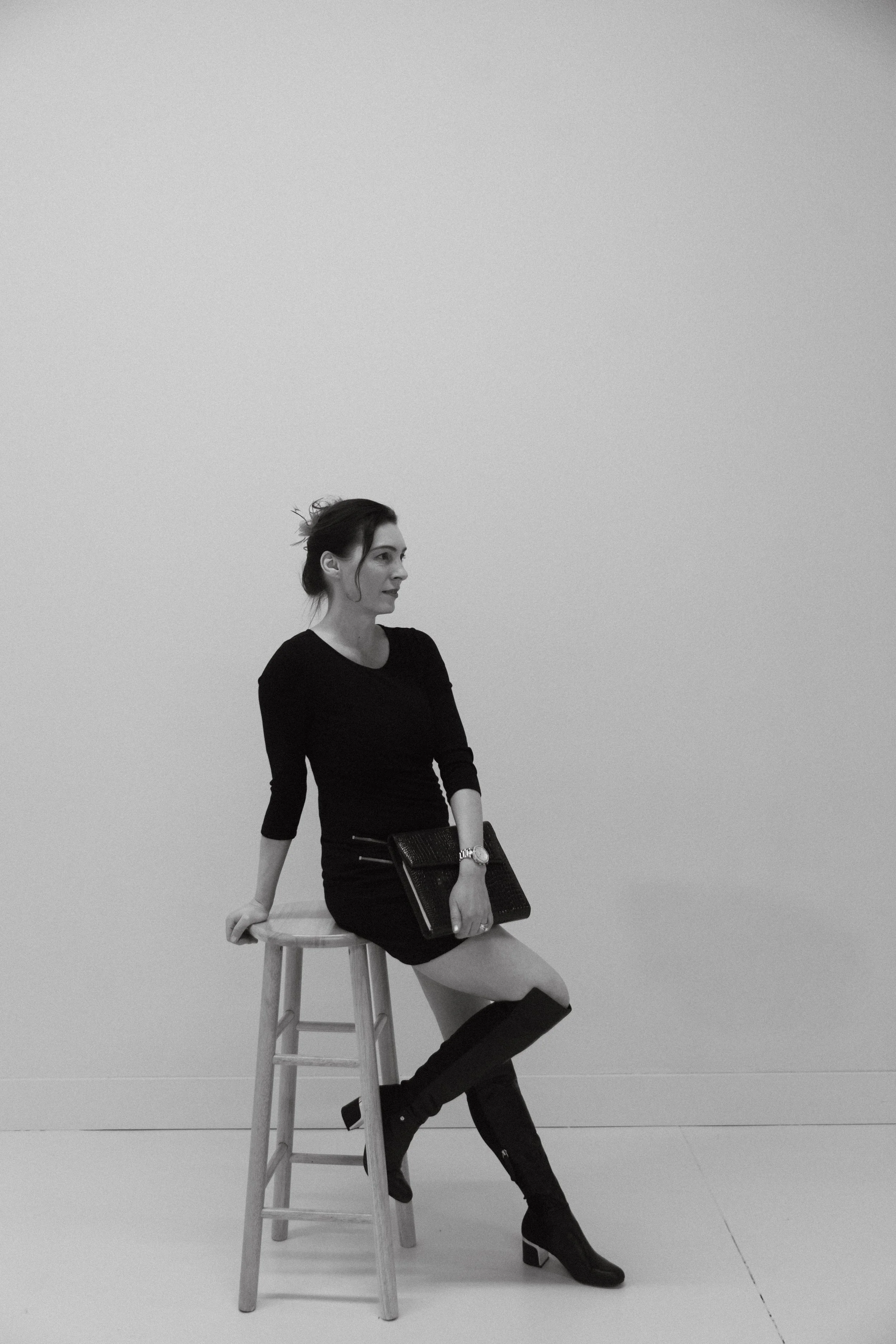 Black and white image of Dr. Kat leaning on a wooden stool, wearing a black dress and boots, holding a pocketbook in front of her lap, one arm leaning on the stool