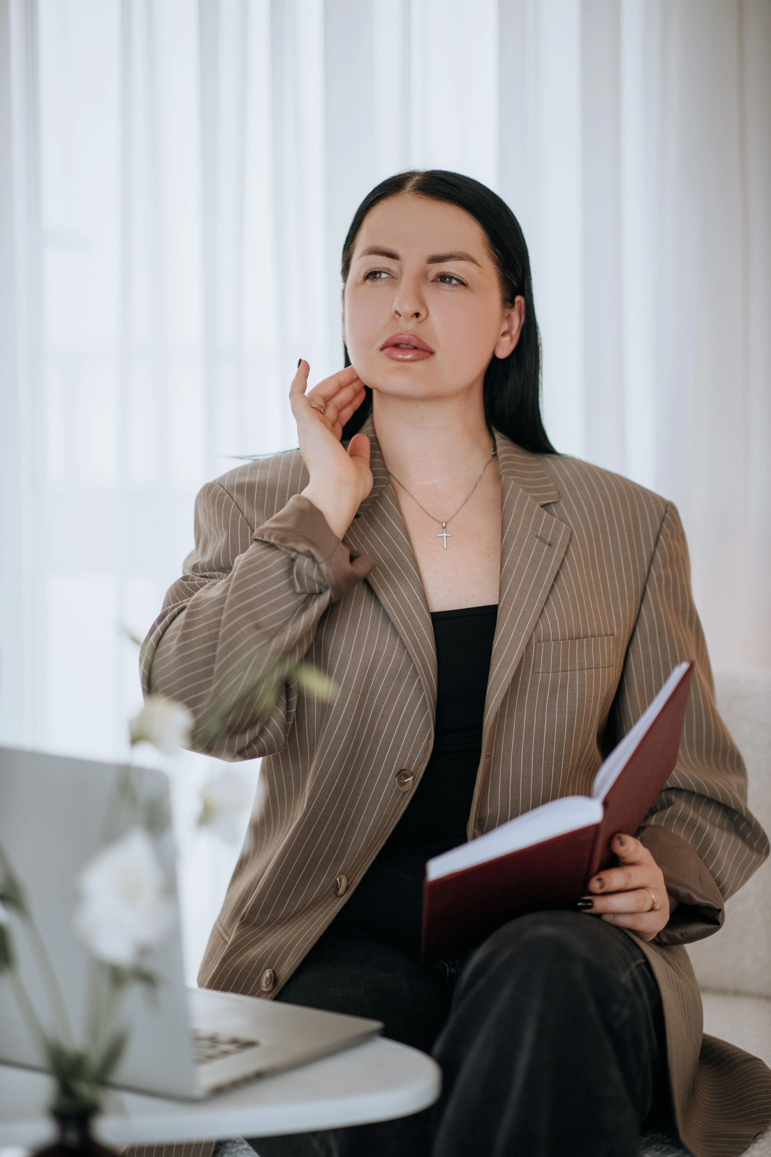 Woman contemplating success in a thoughtful way, sitting by her computer holding a book