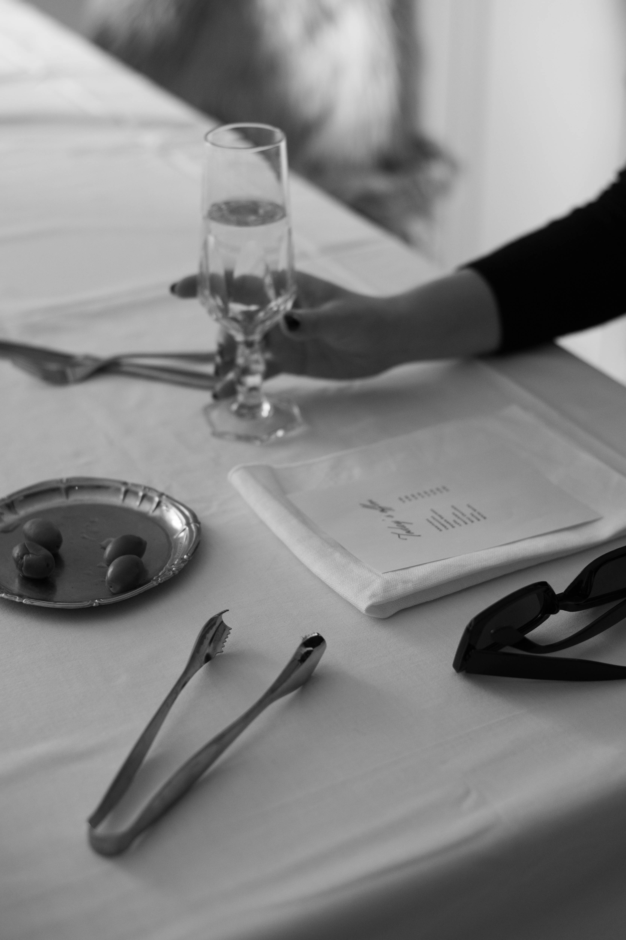a black and white scene of a dinner table with a female hand holding a glass of champagne, with a menu on a napkin in front of her, a small silver tray with grapes and tongs on the white table cloth