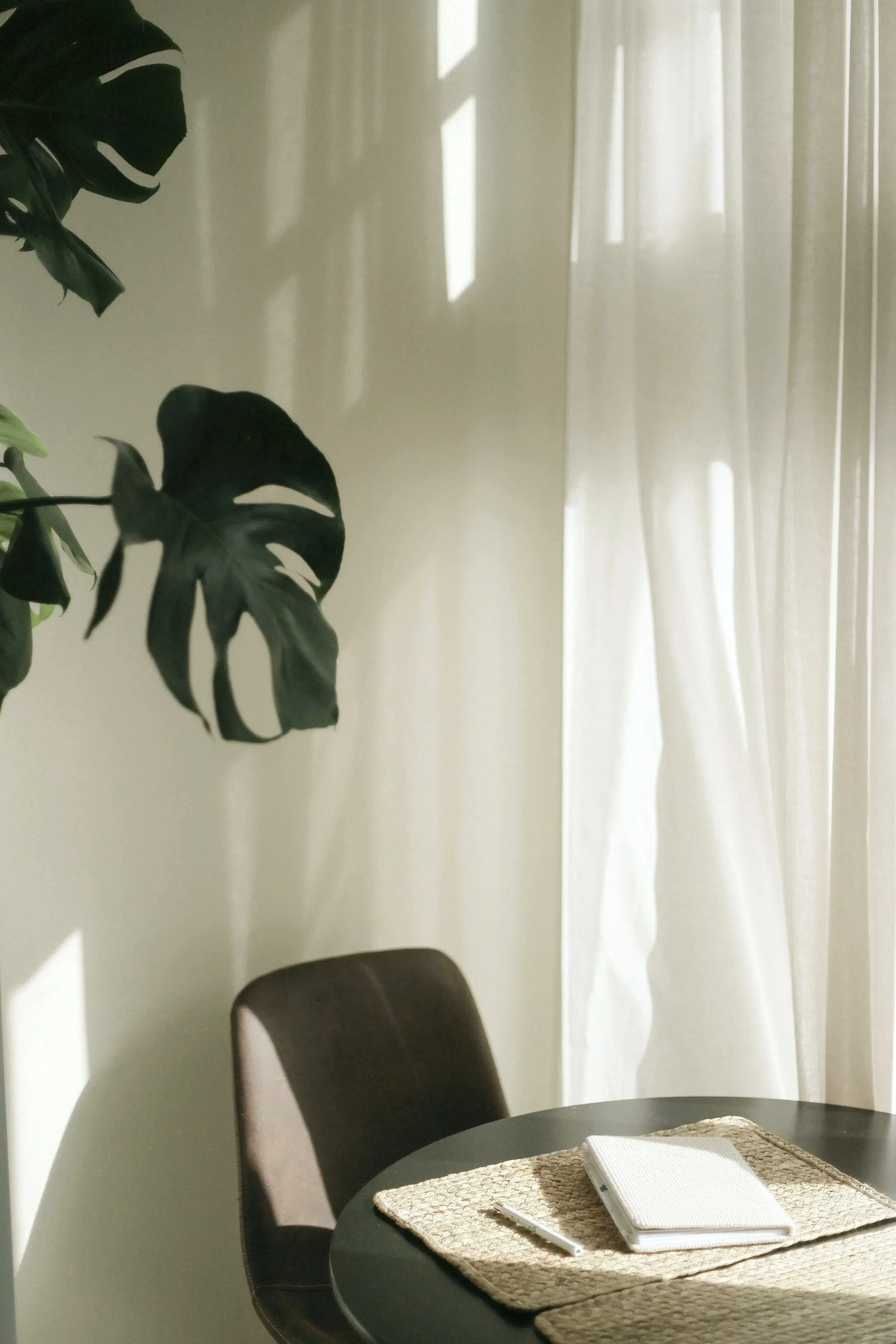 A serene, sunlit corner of a room with a minimalist black table, a brown chair, and a woven placemat. On the placemat rests a white notebook and silver pen.