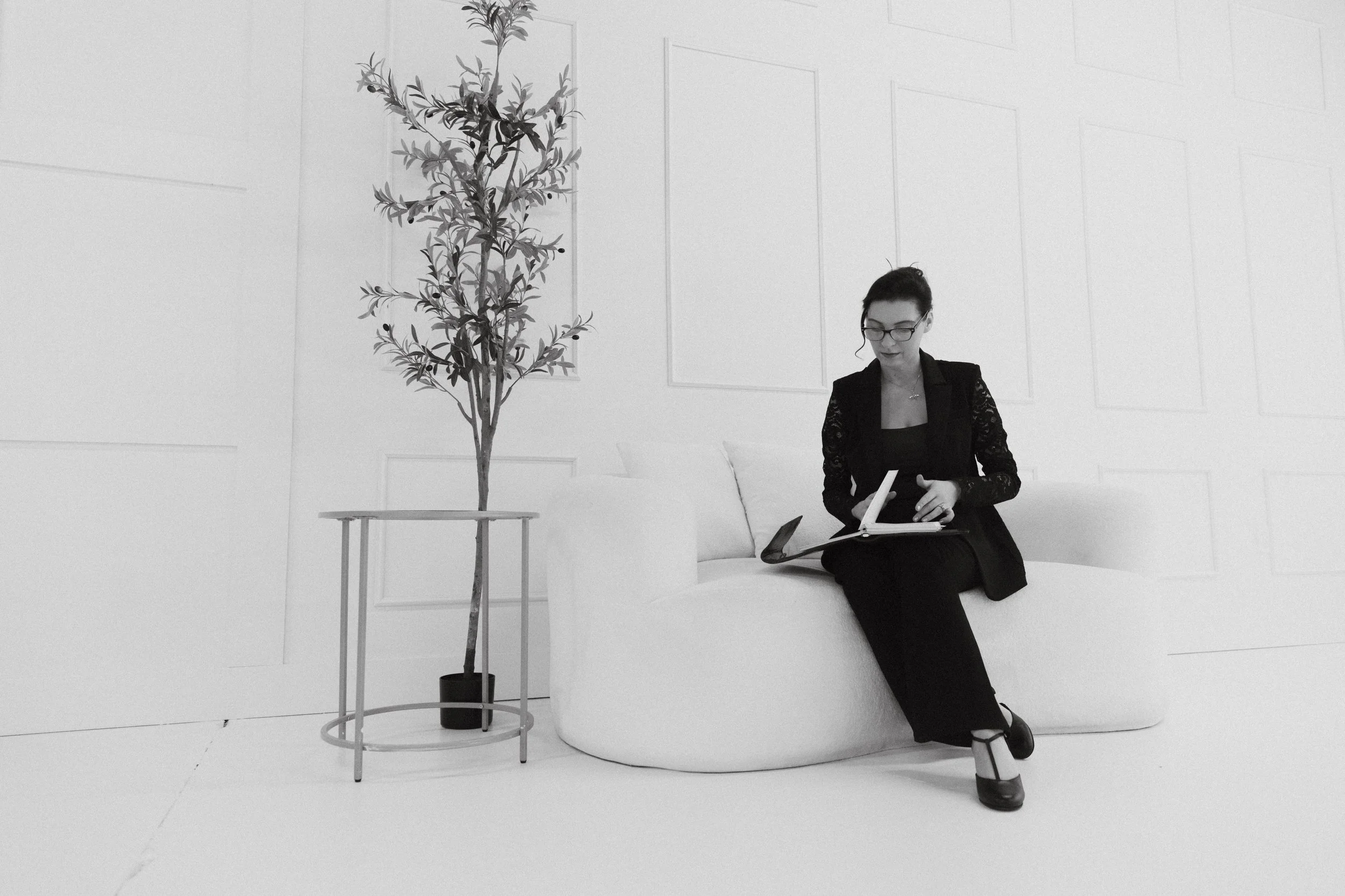 Dr. Kat seated thoughtfully on a white sofa in a minimalist studio space reviewing notes, reflecting Biological Leadership™, nervous system awareness, emotional presence