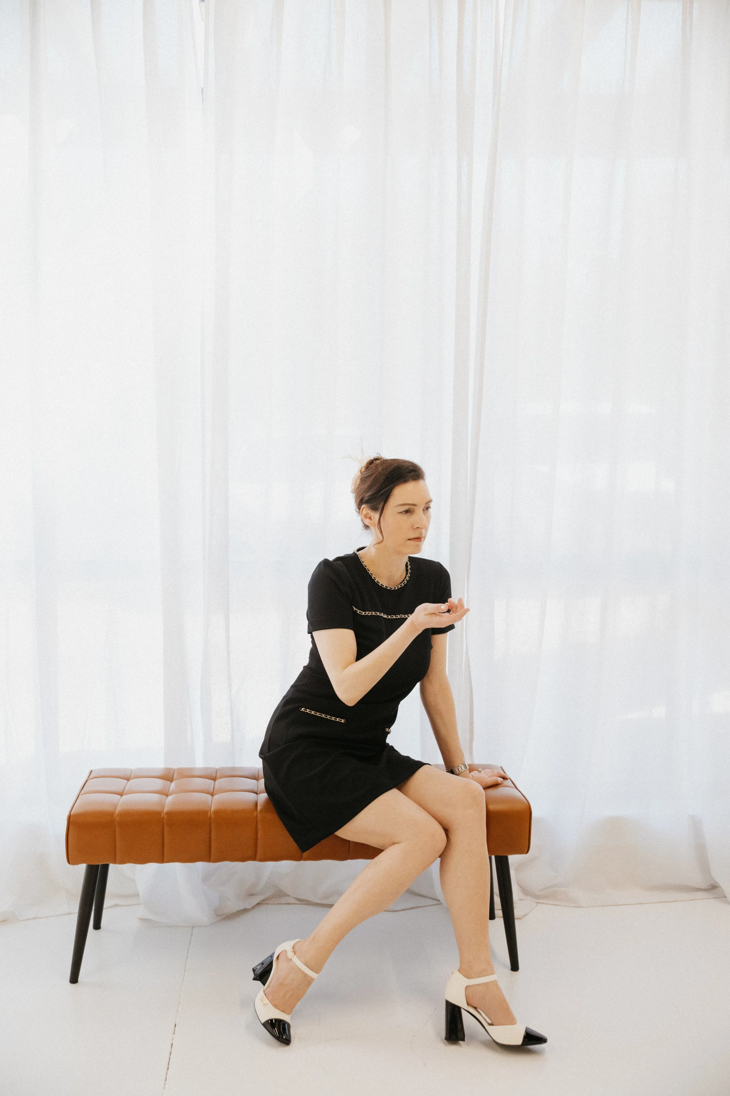 Dr. Kat seated thoughtfully on a leather bench in a softly lit studio space, reflecting emotional presence, self-reflection, and the quiet pressure of navigating growth, responsibility, and personal expansion.