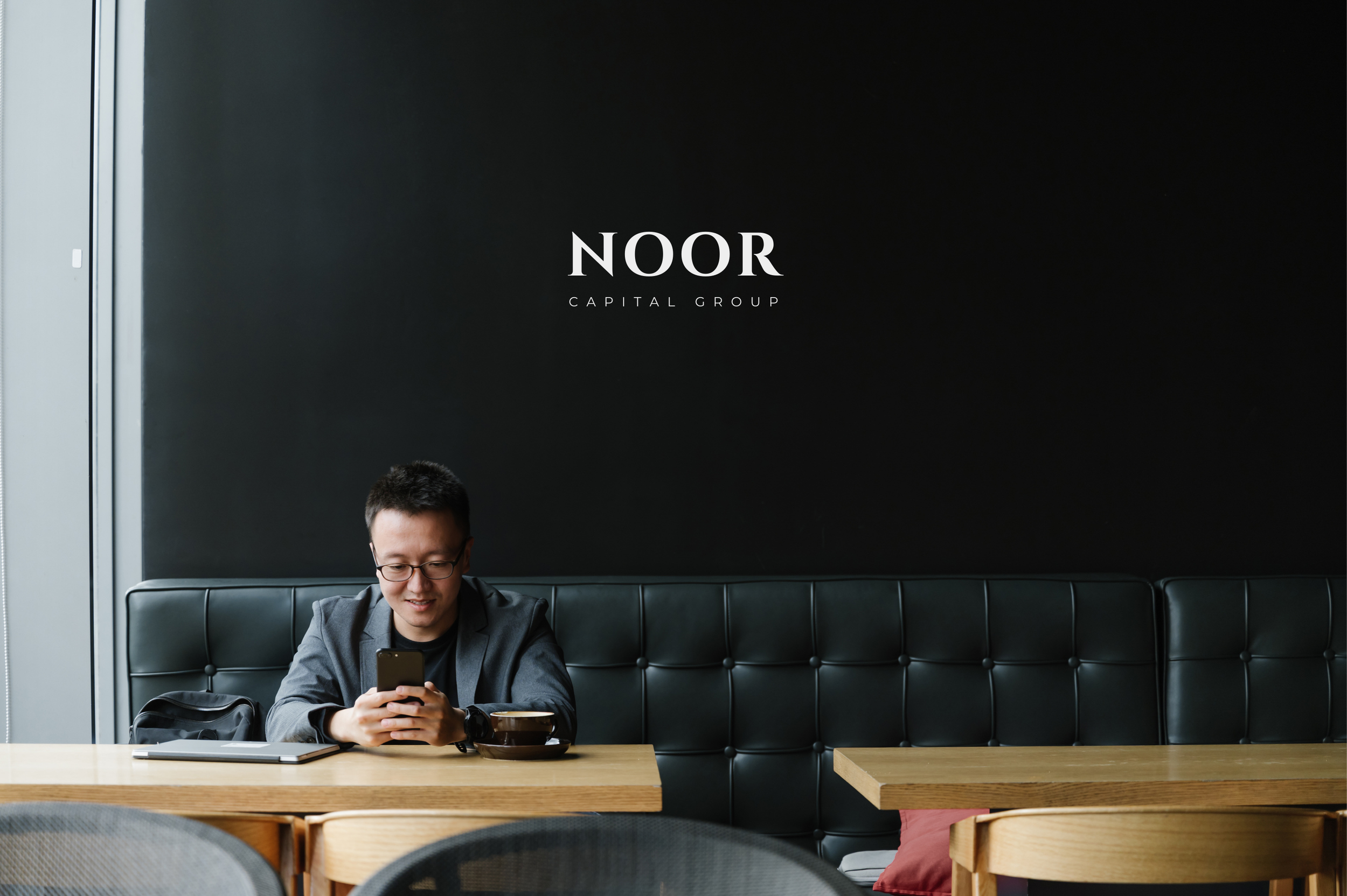 A man with glasses, wearing a dark blazer, sitting at a wooden table in a cafe, using his smartphone. There is a laptop, a black backpack, and a cup of coffee on the table. The background shows a black wall with the words 'NOOR CAPITAL GROUP' written in white.