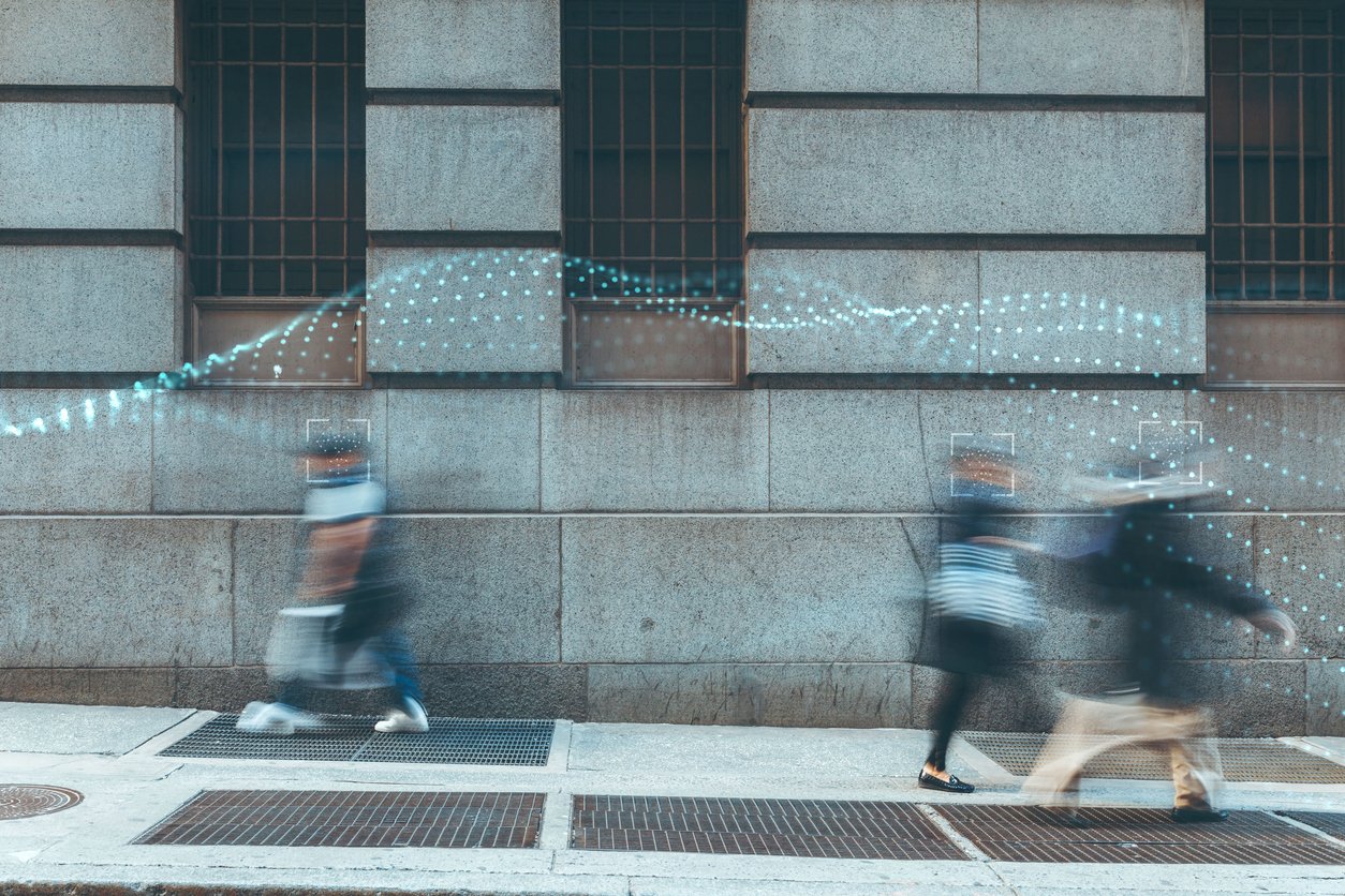 Blurry pedestrians walking on a city sidewalk in front of a concrete building with three windows, with digital trail effects overlaying parts of the scene.