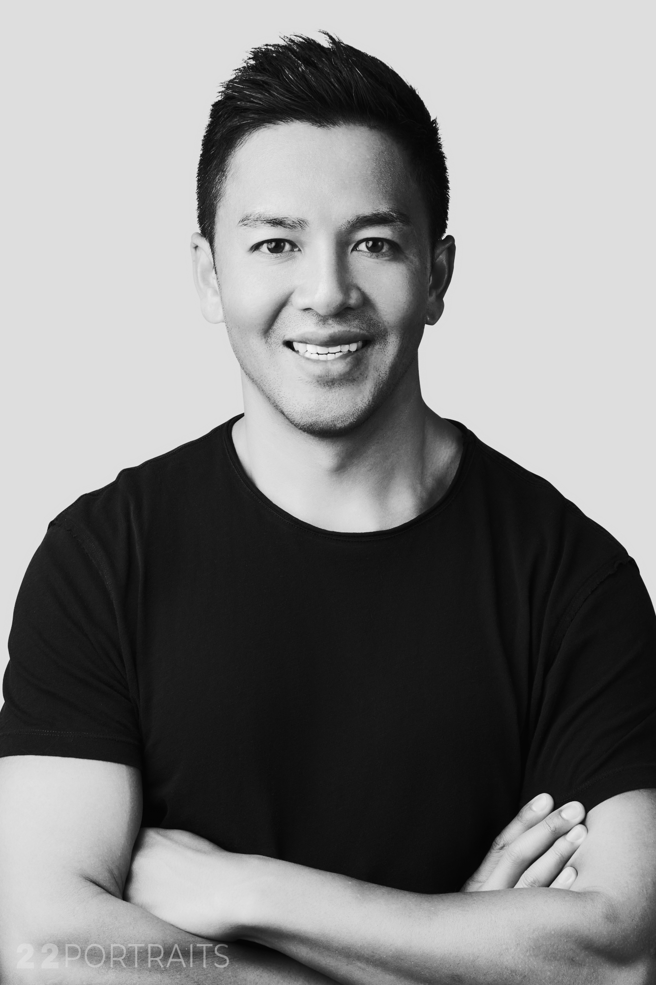 Black and white portrait of a man smiling with crossed arms, wearing a black t-shirt, against a plain light background.