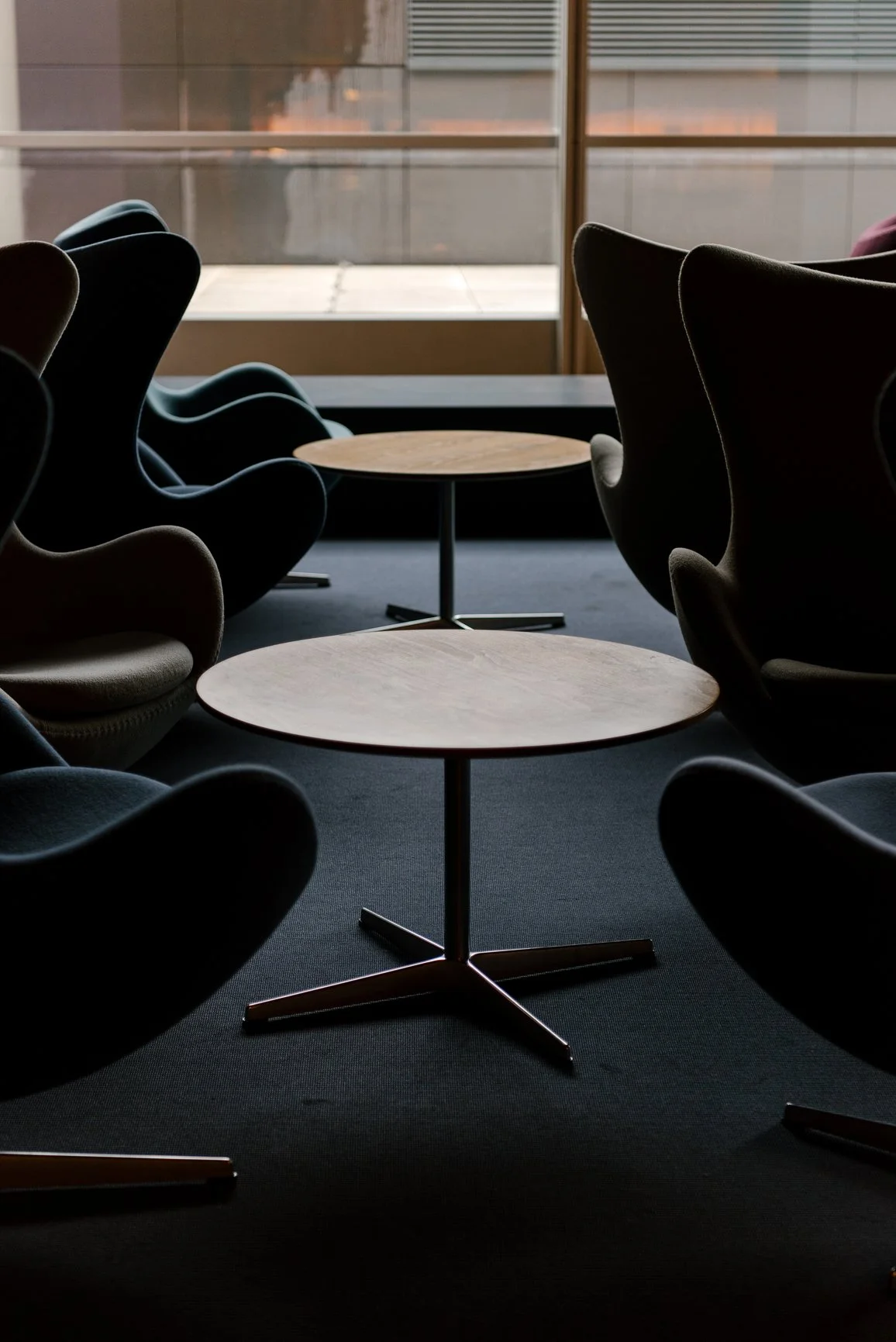 Empty modern lounge with black armchairs, round wooden tables, and large windows, in low light.