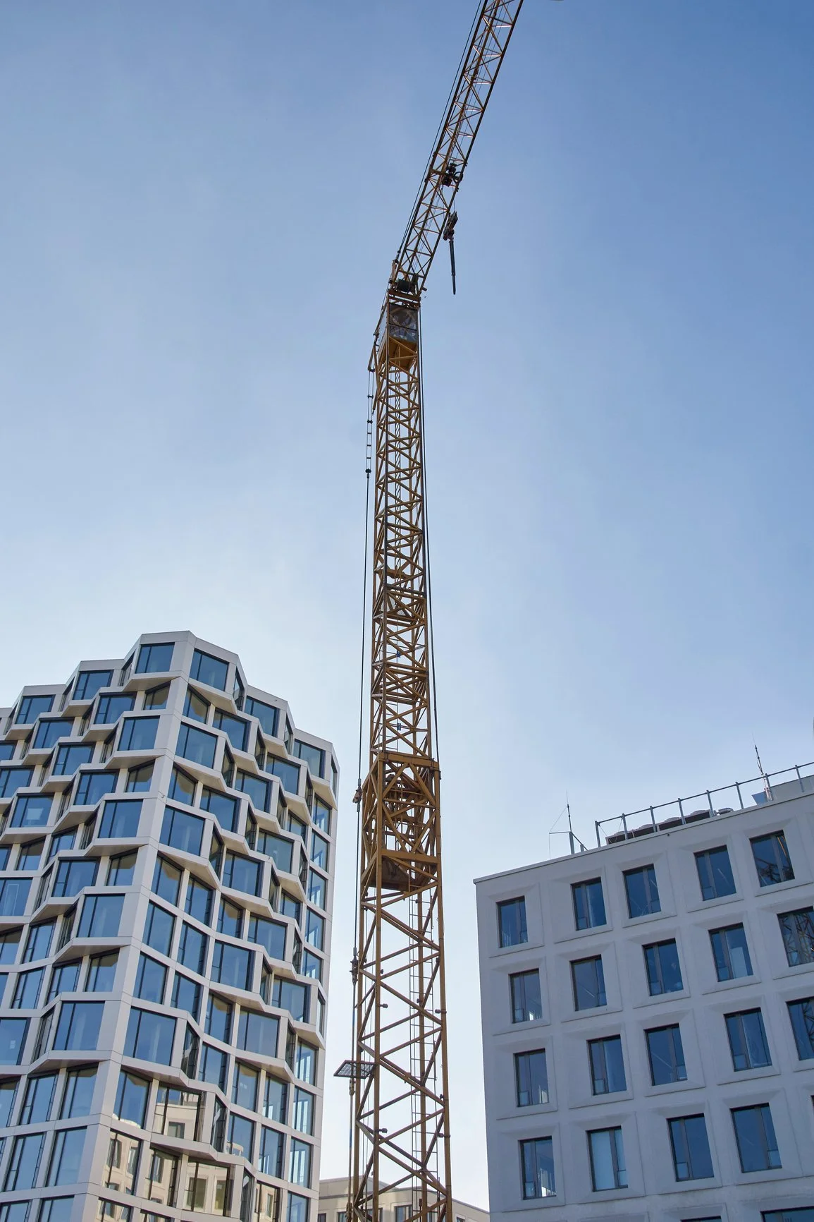 A tall construction crane between two modern multi-story buildings with blue skies in the background.