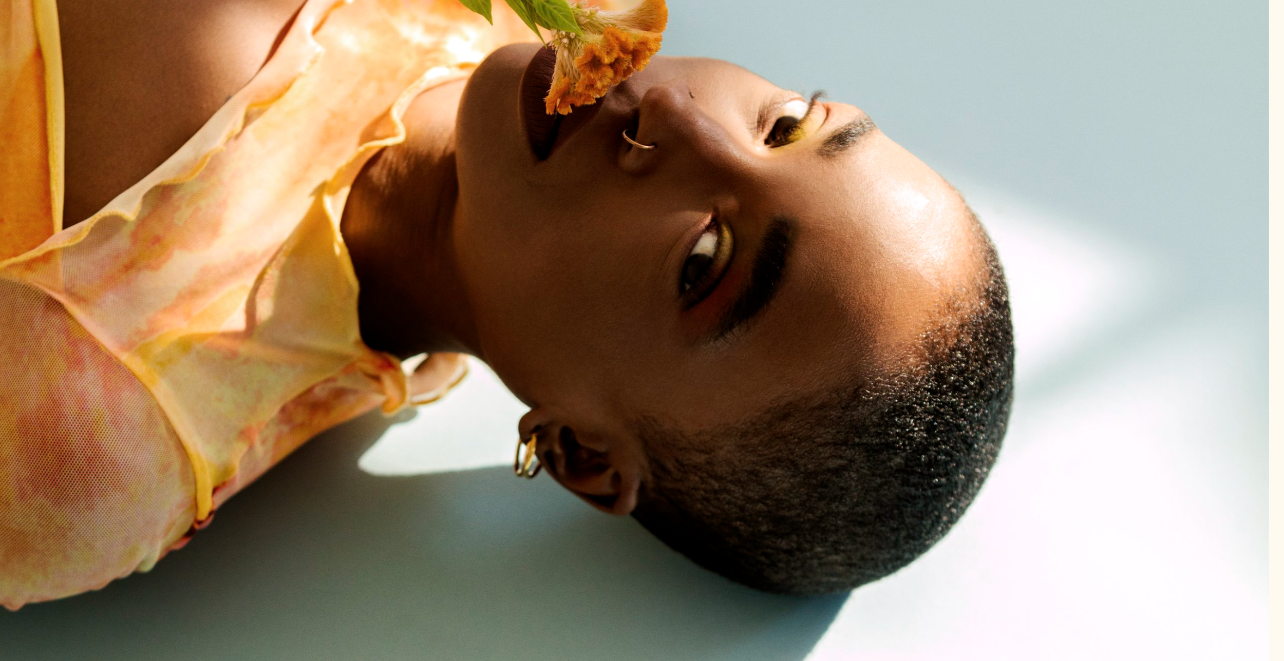 Close-up portrait of a person lying down, with short curly hair, wearing gold hoop earrings, gold eyeshadow, and a sheer yellow top with orange accents, looking directly at the camera.