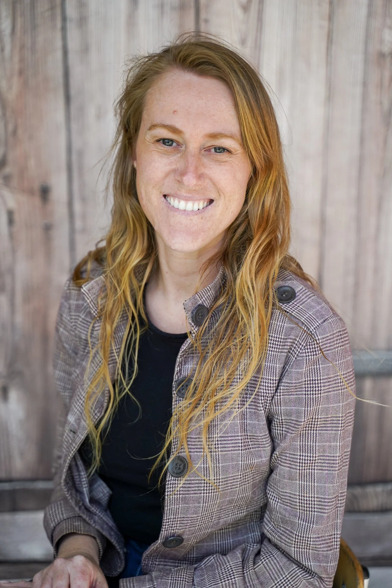 Headshot of a smiling woman with red hair, wearing a checkered blazer, sitting in front of a wooden background.