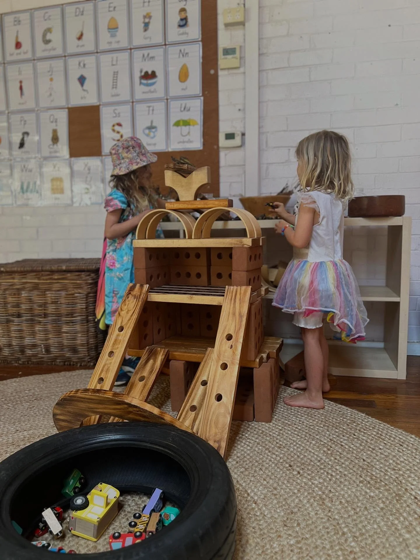 Two young girls playing with wooden and toy cars in a room with a wall display of illustrated alphabet cards.