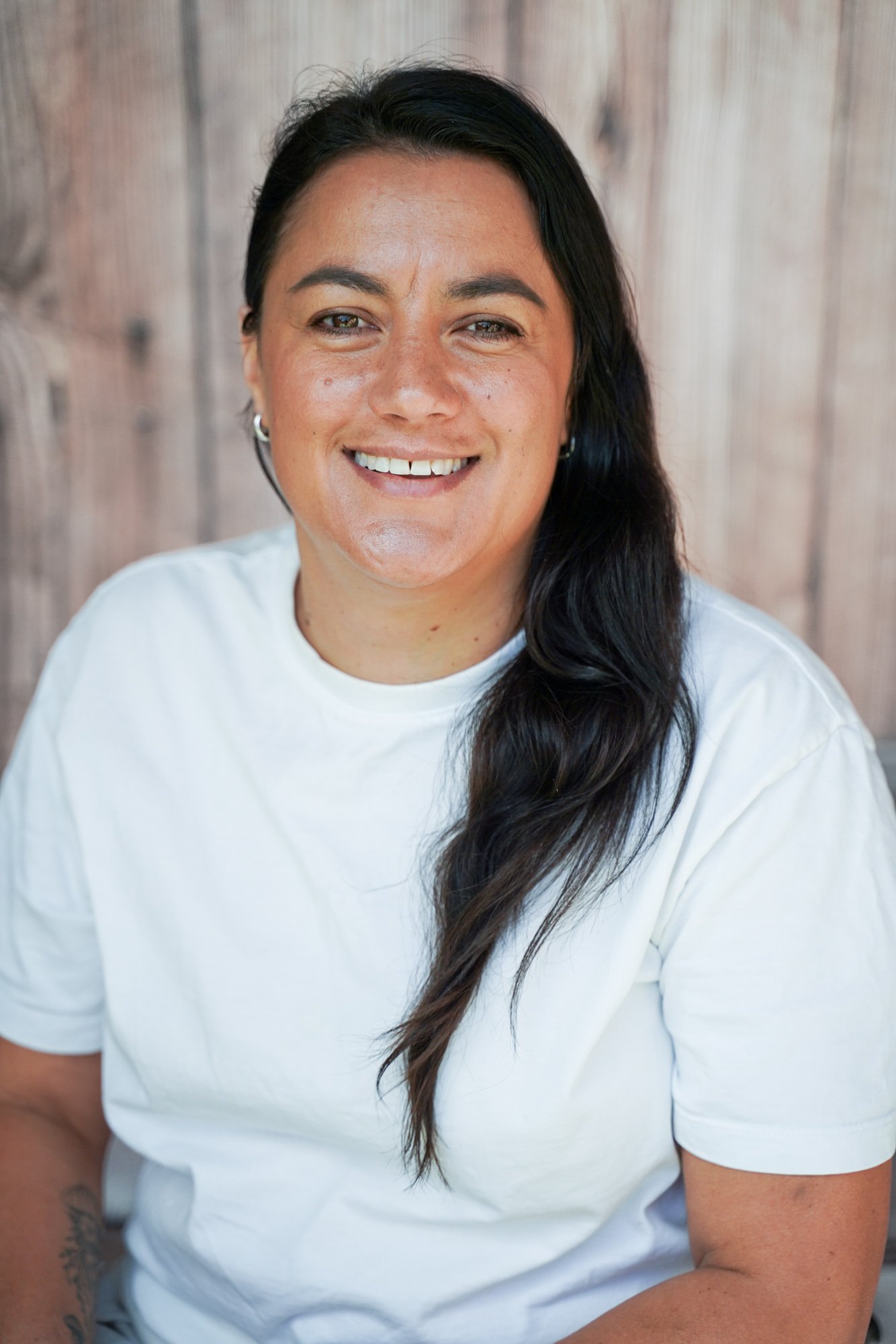 Close-up of a woman with long dark hair, wearing a white t-shirt, smiling in front of a wooden background.
