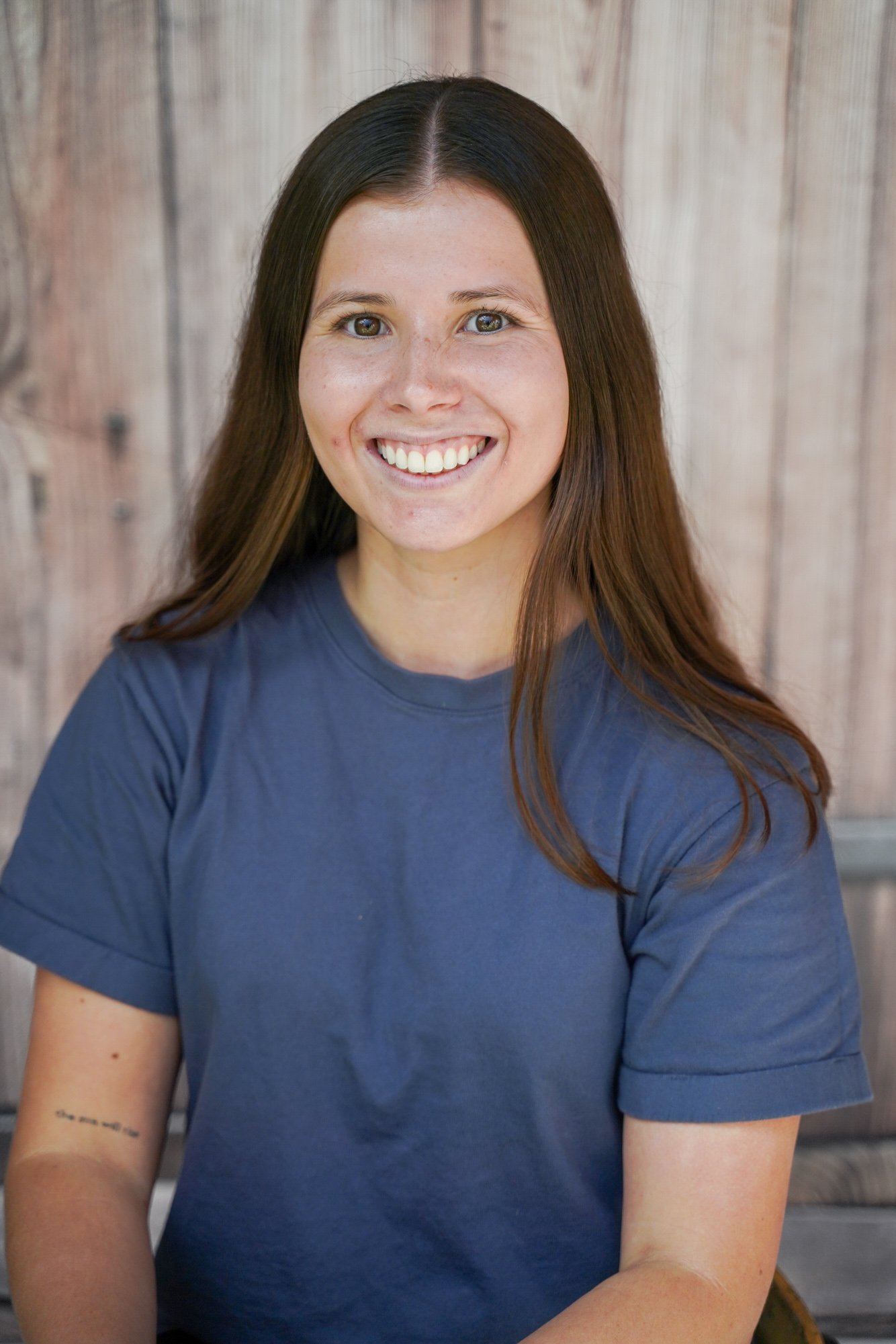 A young woman with long brown hair, smiling in front of a wooden wall, wearing a blue T-shirt.