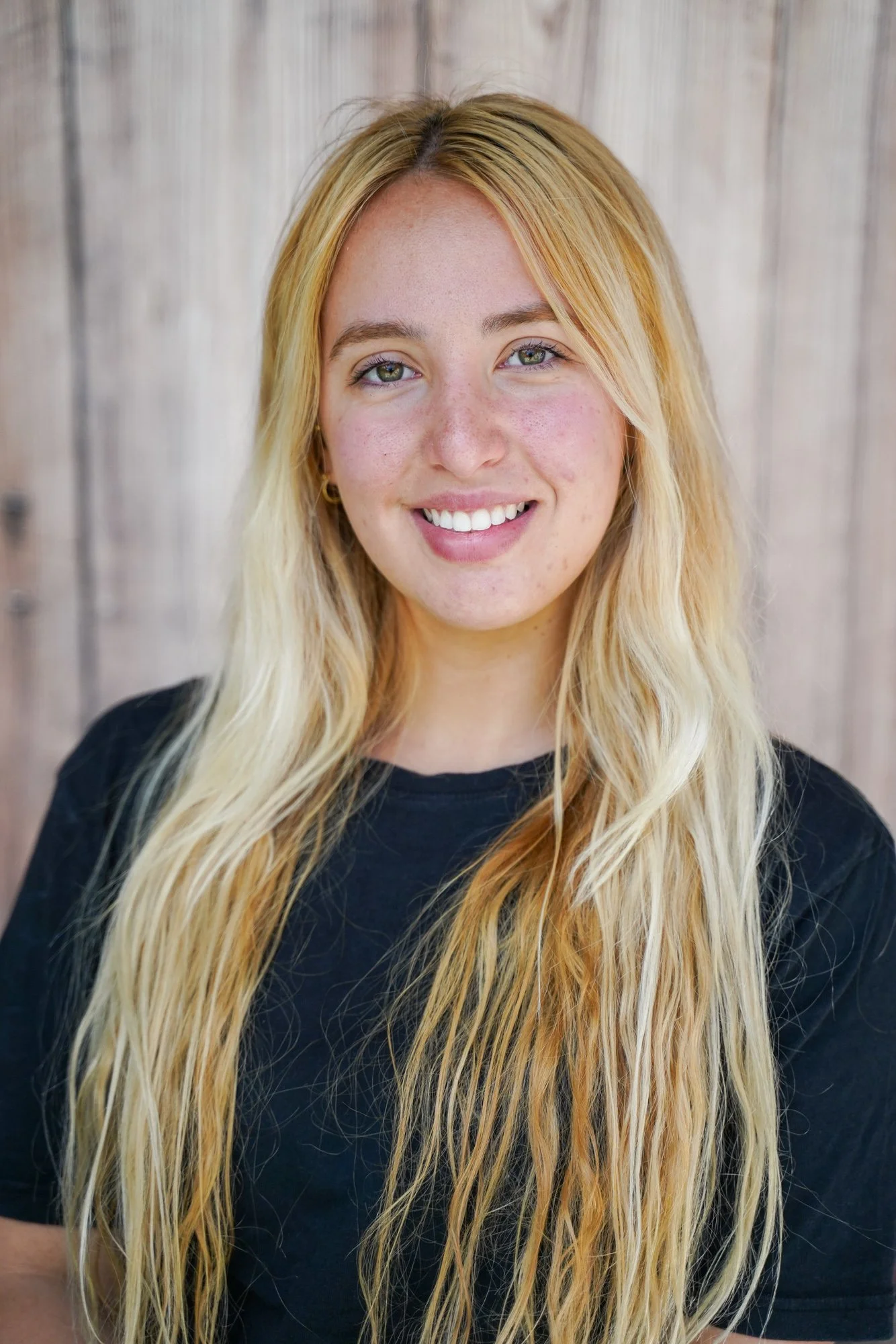 A young woman with long blonde hair and green eyes smiling in front of a wooden background.