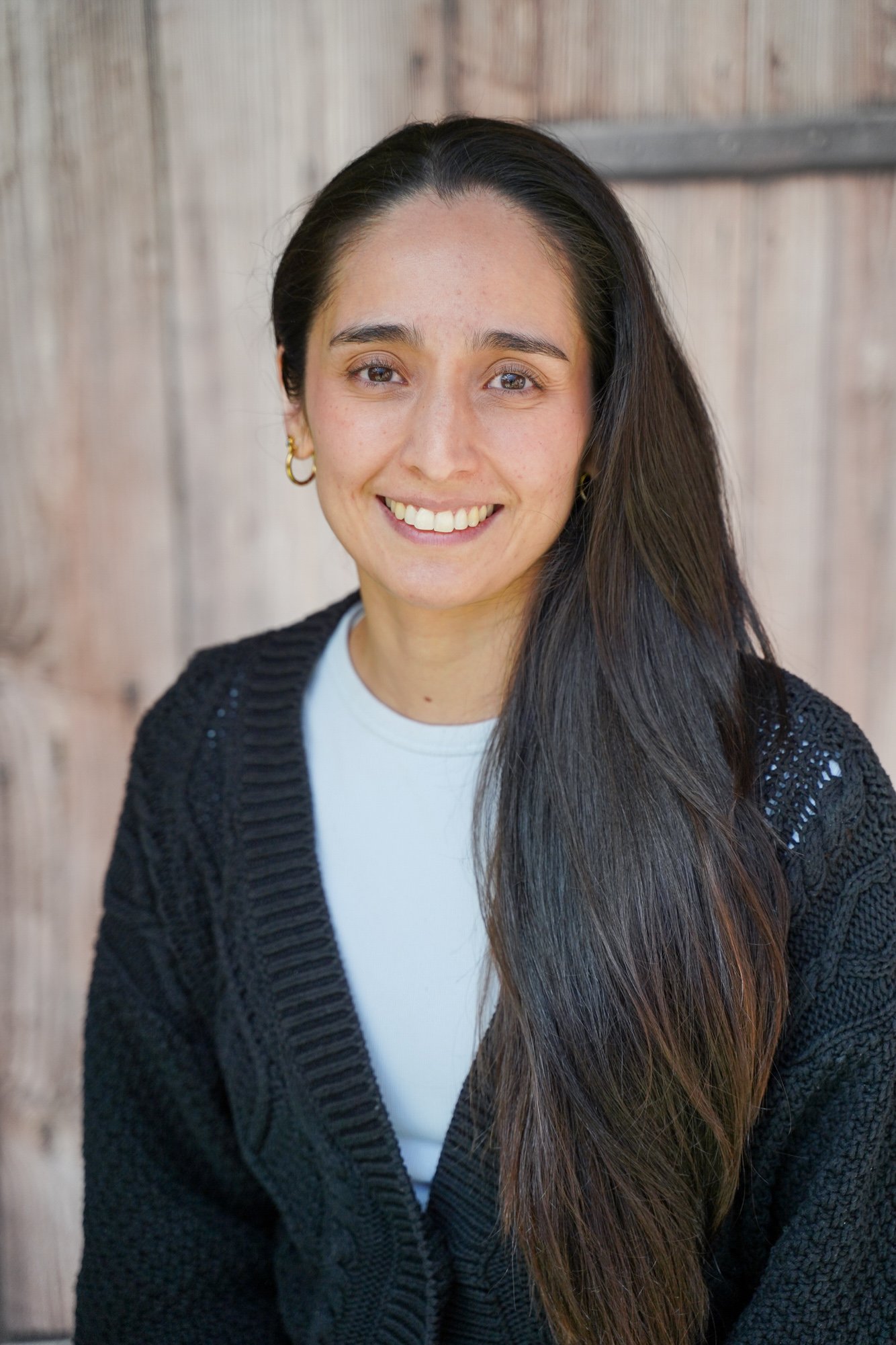 A portrait of a woman with long, dark hair, smiling, wearing a black cardigan and gold hoop earrings, standing in front of wooden background.