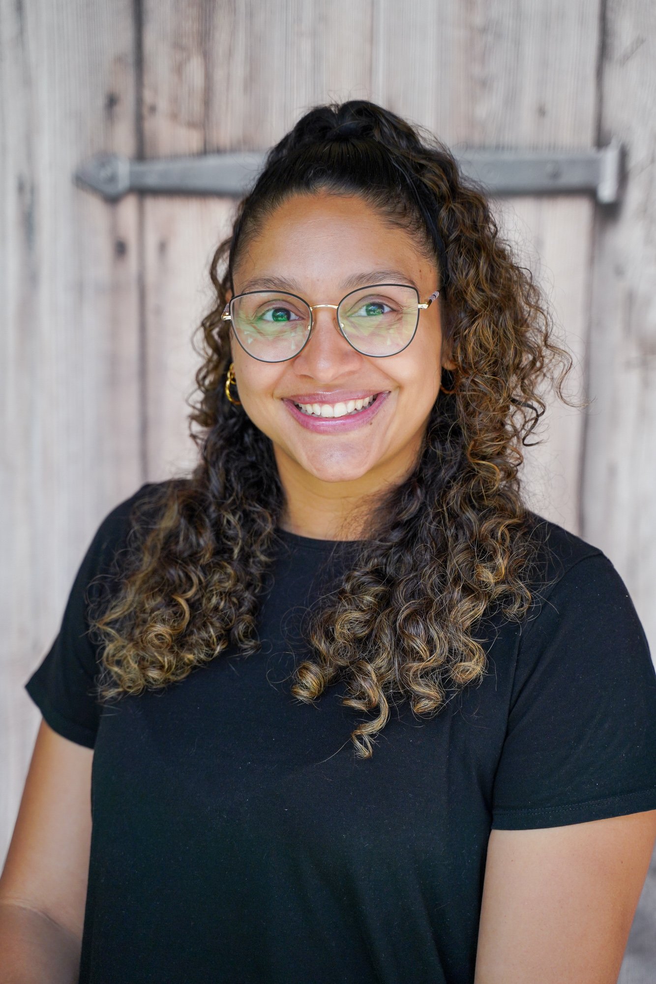 A woman with curly hair, wearing glasses and a black t-shirt, smiling in front of a wooden background.
