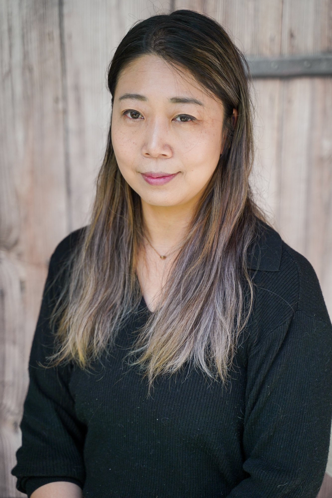 A woman with light skin and long, wavy hair with dark roots and lighter tips, wearing a black top, sitting in front of a wooden fence.