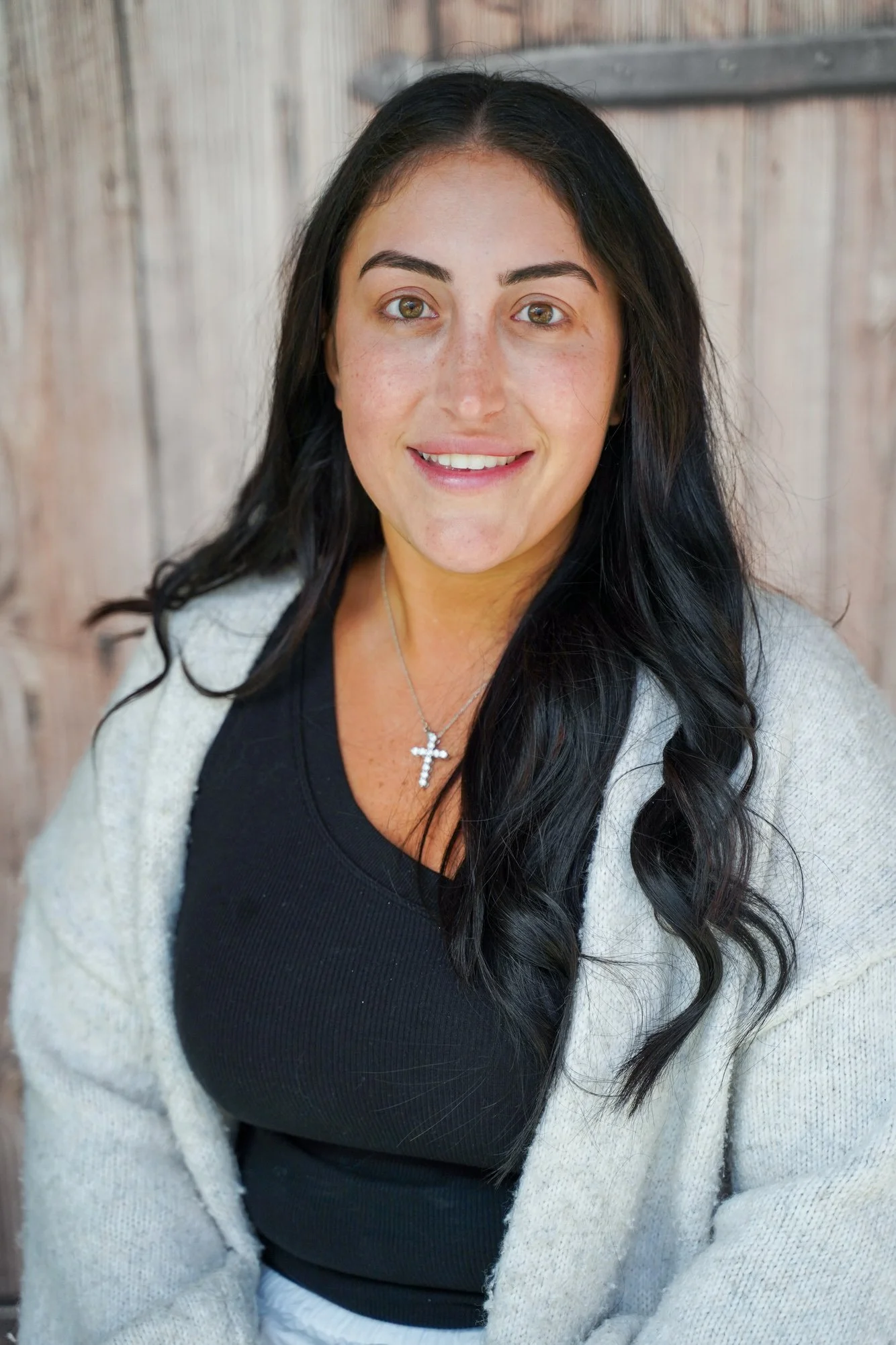 A woman with long dark hair, wearing a black shirt and a light-colored cardigan, smiling, standing in front of a wooden background.