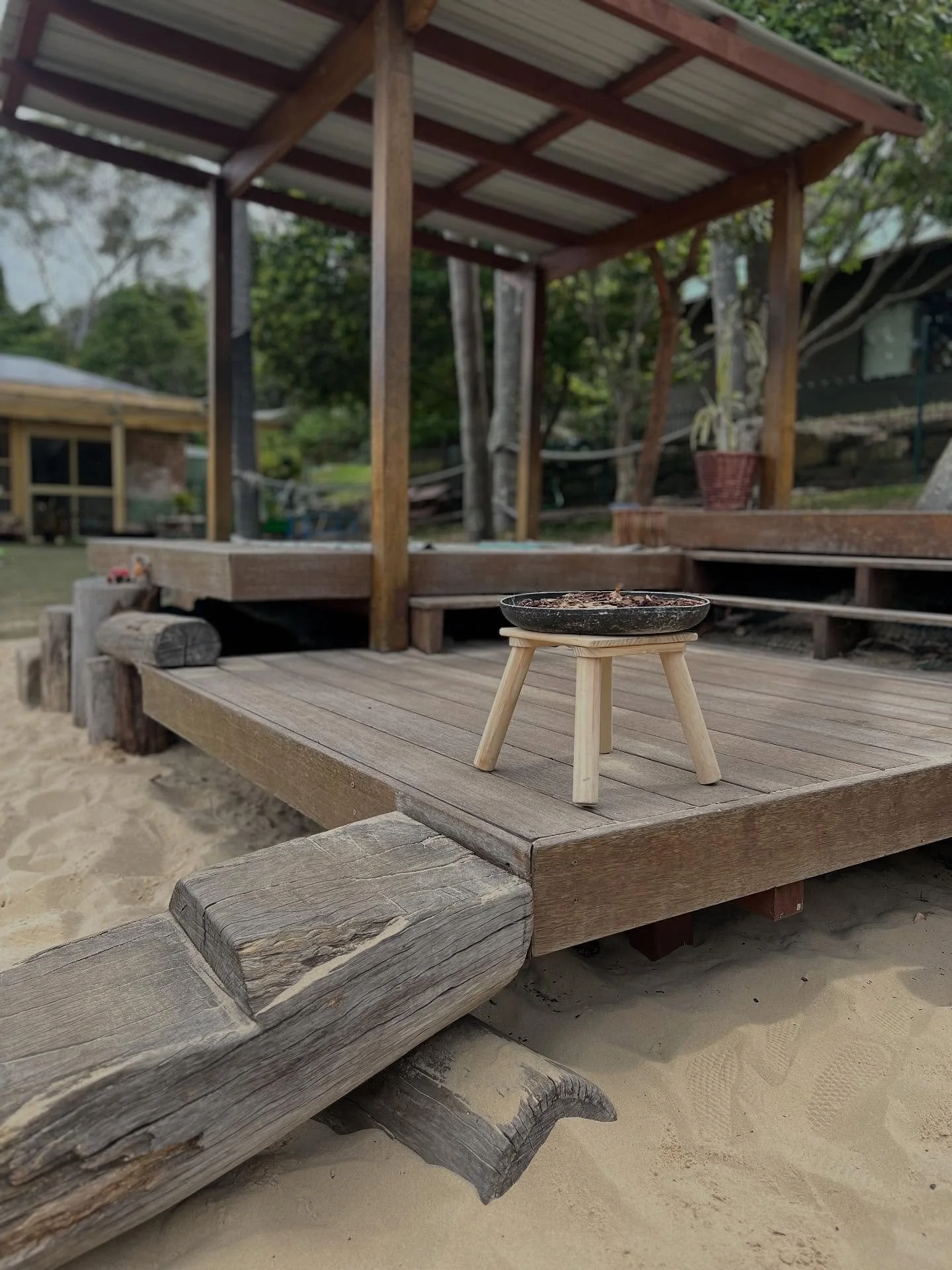 A small wooden stool on a wooden deck with a black ashtray filled with cigarette ash and small debris, overlooking sandy ground and an outdoor structure in the background.