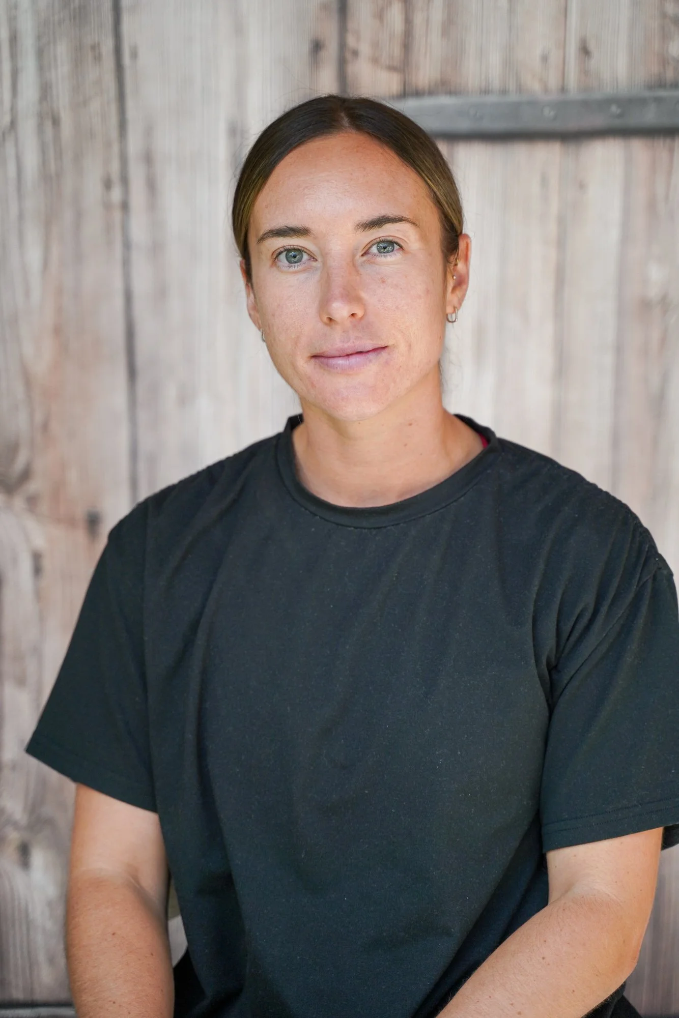 A woman with short brown hair, light skin, and blue eyes, wearing a black T-shirt, sitting in front of a wooden background.
