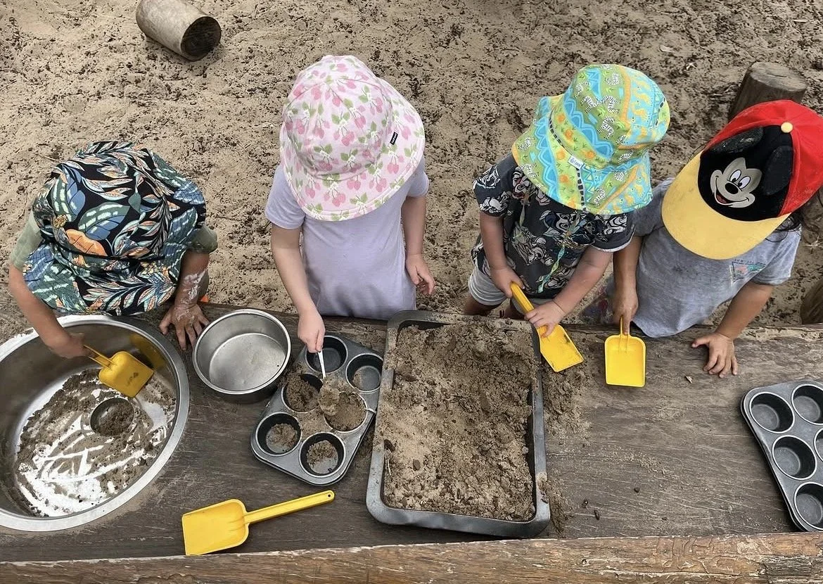 Four young children wearing hats and casual clothing are playing with sand and small shovels in an outdoor sandbox, with various metal baking tins and a tray for making sand cupcakes.
