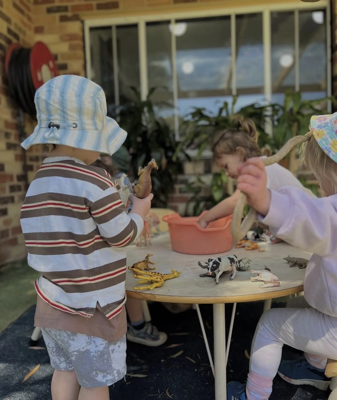 Children playing with toy animals at a table outdoors, some wearing hats, with a brick wall and window in the background.