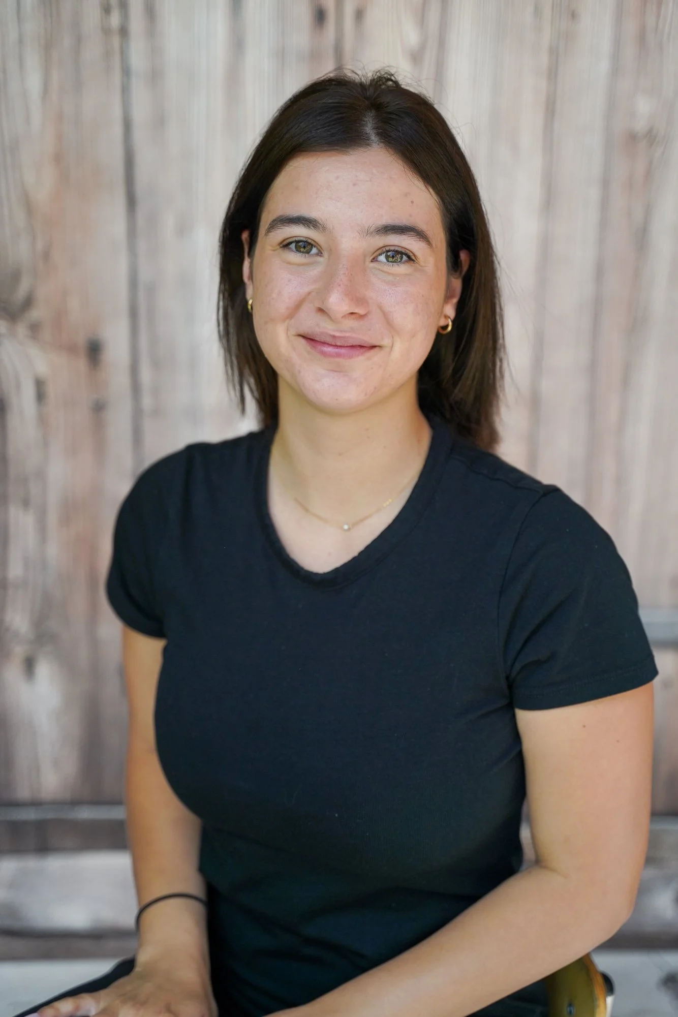 A young woman with short dark hair and light skin smiling, wearing gold hoop earrings, a black t-shirt, sitting in front of a weathered wooden wall.