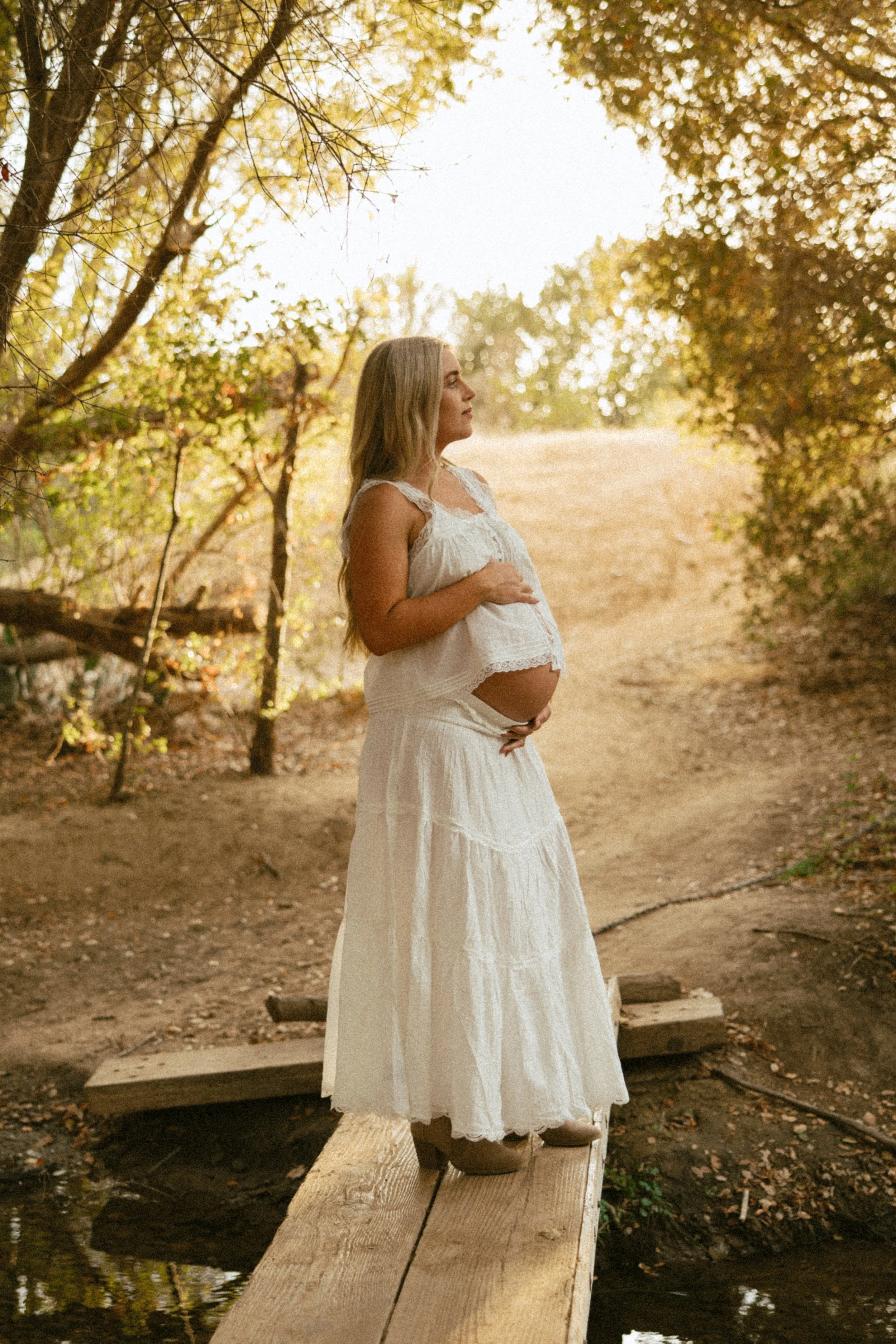 A pregnant woman wearing a white dress and standing on a wooden bridge in a forest with green and orange leaves.